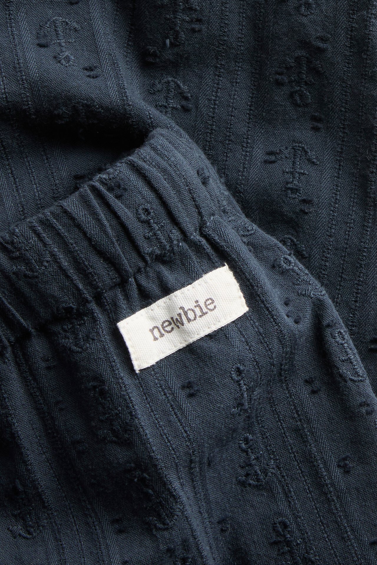 Close-up of dark blue woven cotton pants with an elastic waistband, subtle anchor pattern, and a 'Newbie' tag.