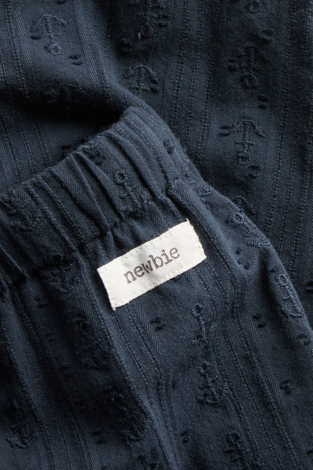 Close-up of dark blue woven cotton pants with an elastic waistband, subtle anchor pattern, and a 'Newbie' tag.
