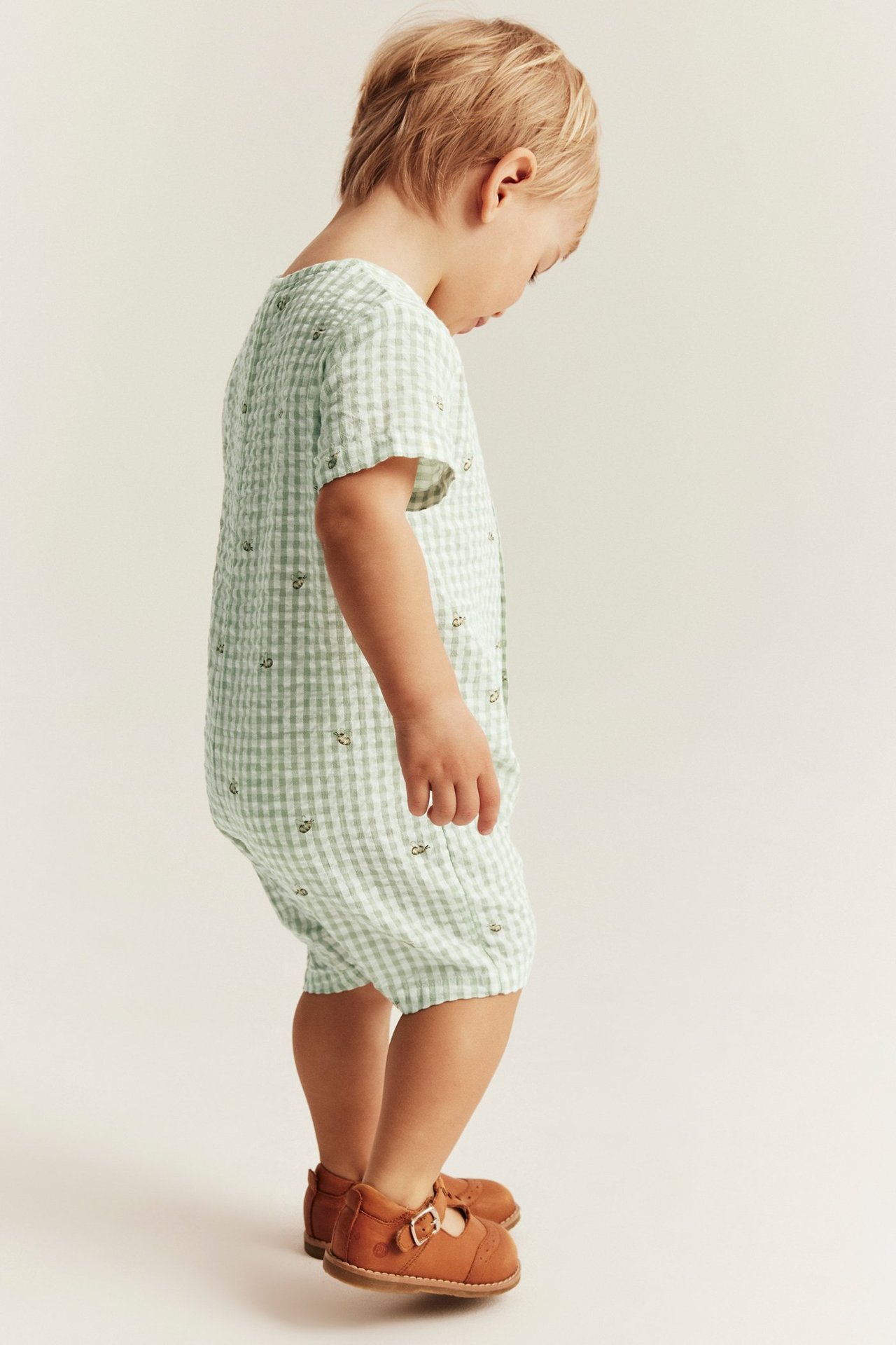 Seitenansicht eines Kindes in grün kariertem Baumwoll-Strampler mit Bienenmuster und braunen Schnallenschuhen.