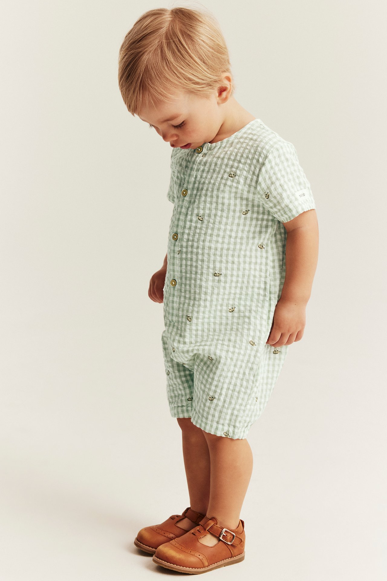 Side view of a baby in a green checkered cotton romper with bee pattern, front buttons, and brown shoes.