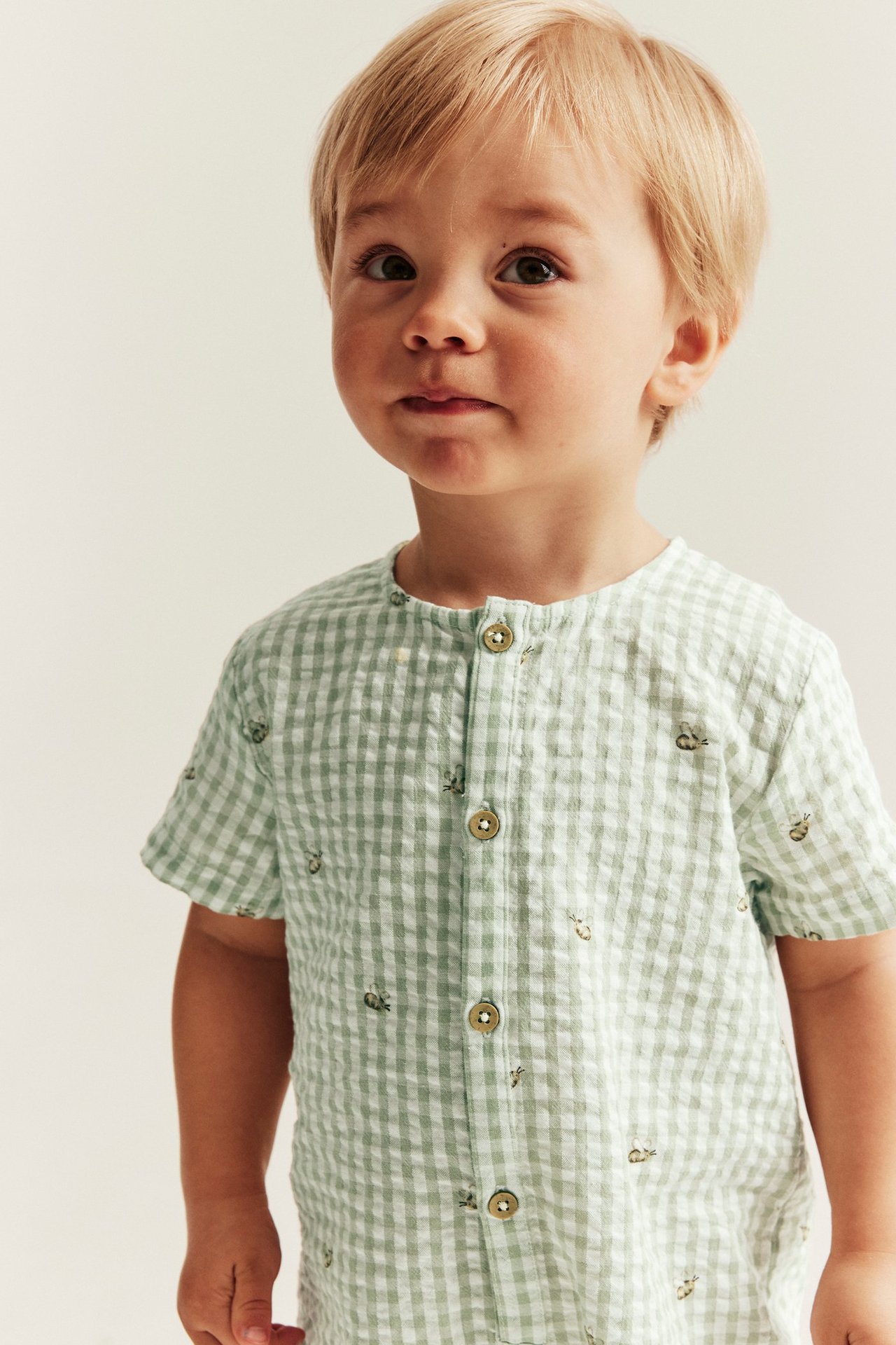 Vorderansicht eines Babys in einem grün-weiß karierten Baumwoll-Overall mit Bienenmuster und Knöpfen.