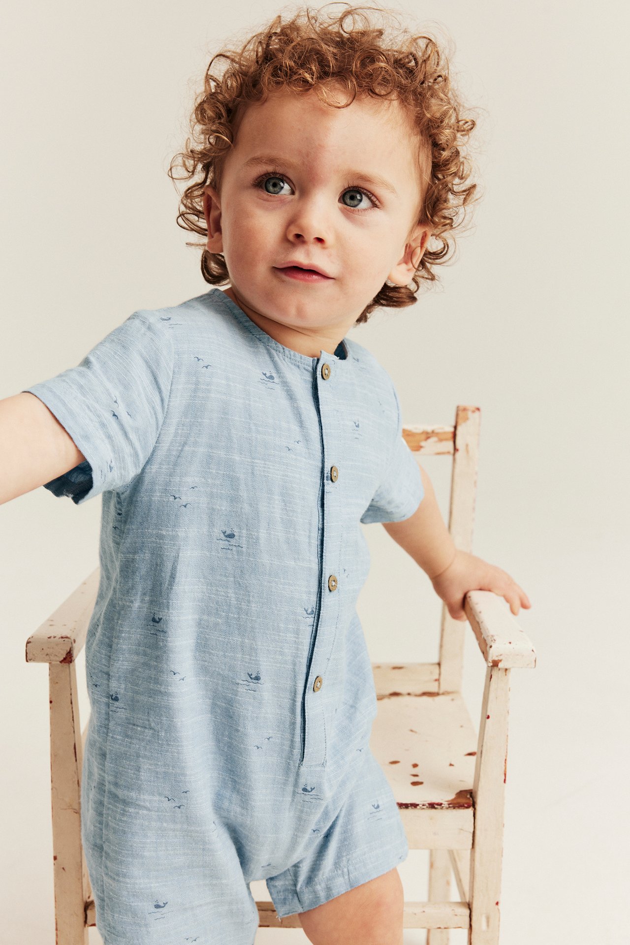 Front-side view of a baby wearing a light blue short-sleeved cotton jumpsuit with whale and bird patterns, featuring front buttons.
