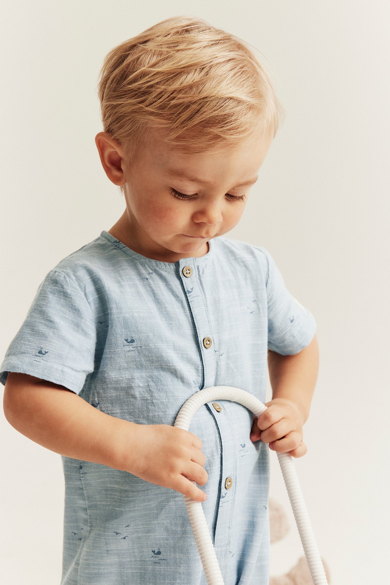 Front view of a baby boy in a light blue, whale-patterned cotton romper with front buttons, holding a white toy.