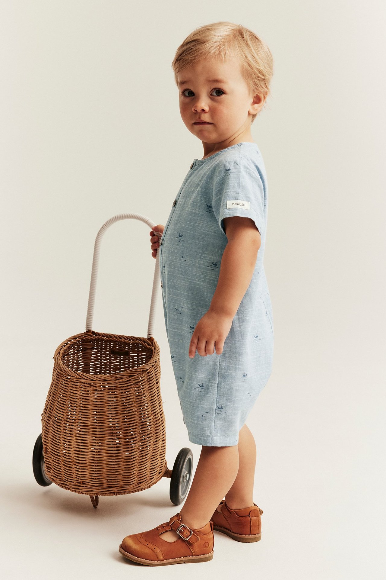 Seitenansicht: Baby in hellblauem Walmuster-Jumpsuit, braunen Schuhen, schiebt Weidenkorb auf Rädern.