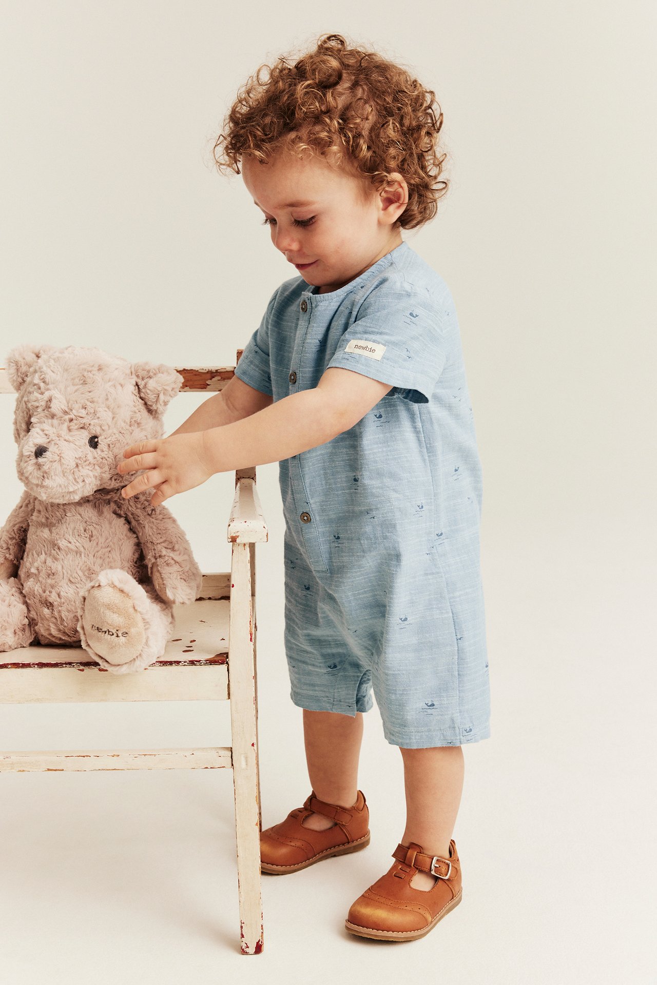 Front view: Baby boy in a light blue cotton whale-patterned short-sleeved romper, brown shoes, touching a teddy bear.
