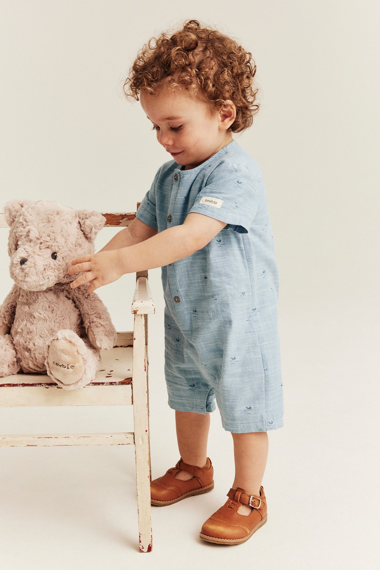 Front view: Baby boy in a light blue cotton whale-patterned short-sleeved romper, brown shoes, touching a teddy bear.