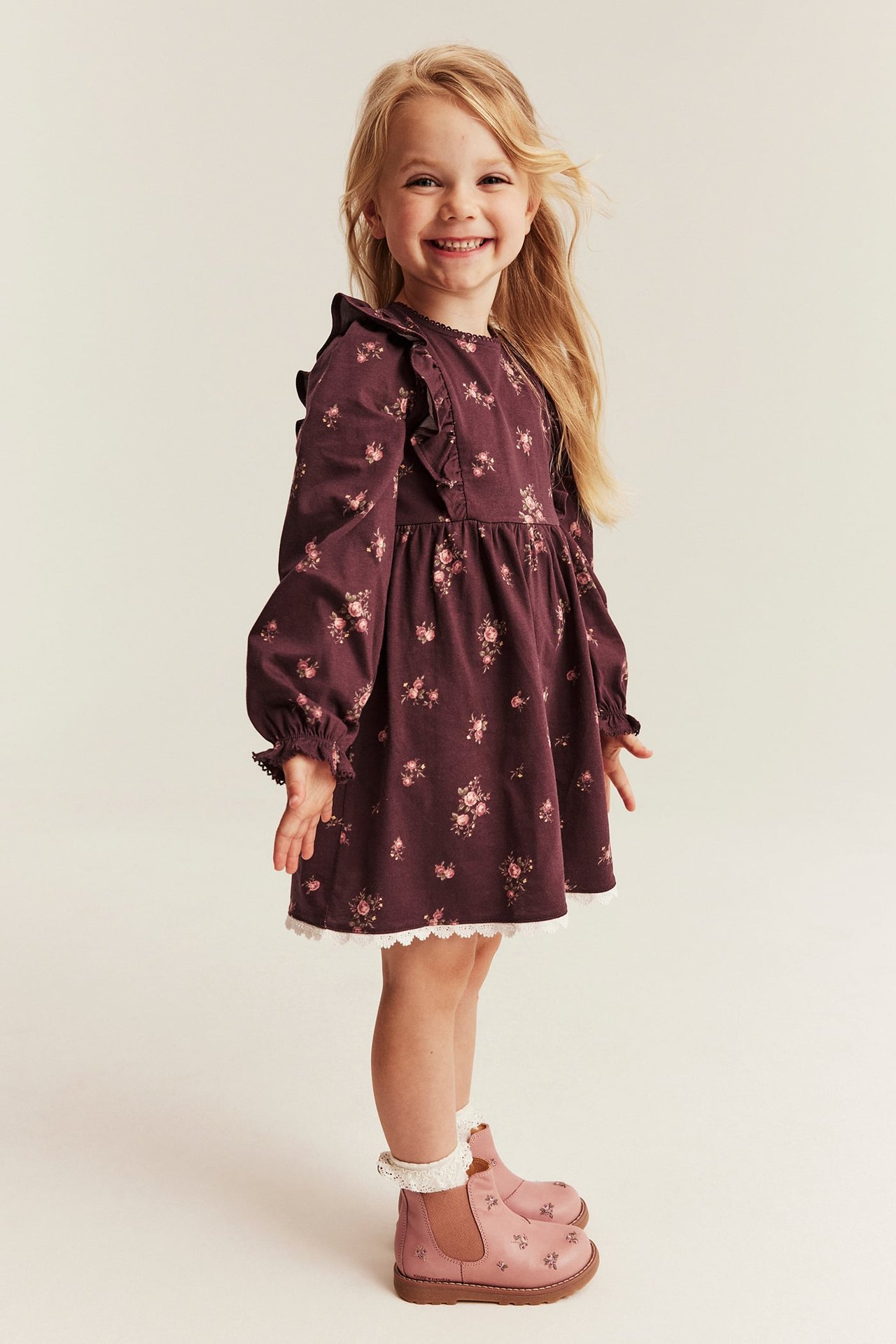 Front view: Kid in dark purple floral ruffled dress with lace, white socks, and pink floral boots.