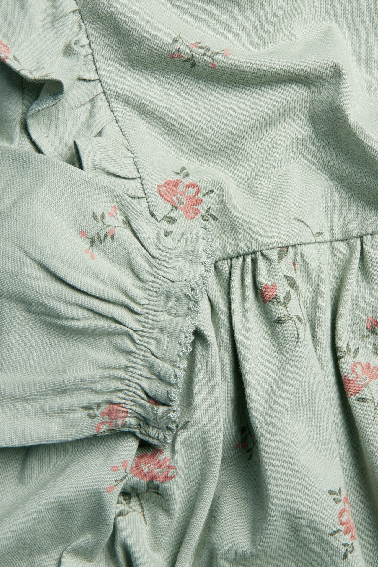 Close-up of a light green floral jersey dress with pink flower print, ruffles, and elasticated picot-edge cuffs.