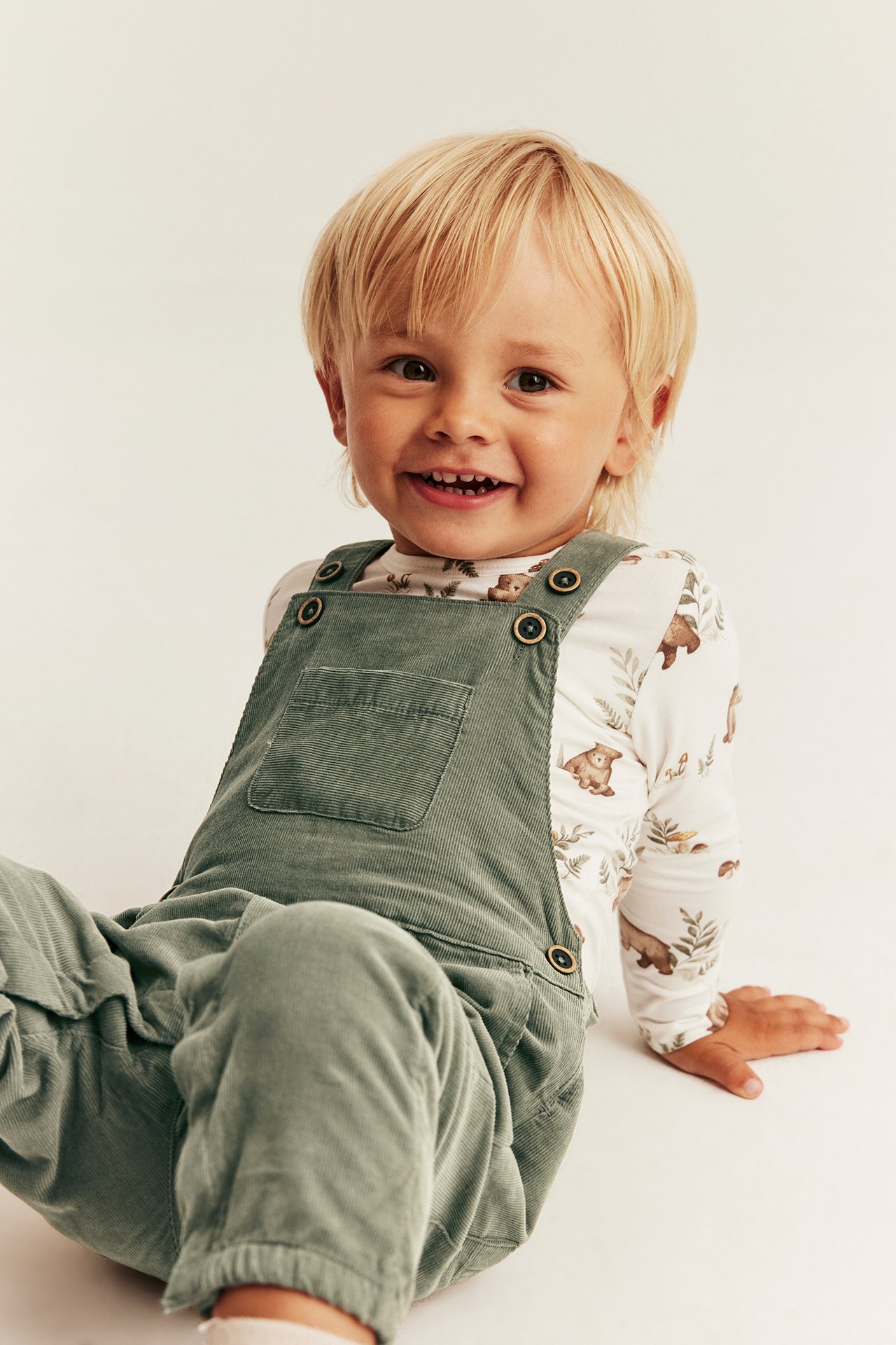 Kind in grüner Cord-Latzhose über weißem Langarmshirt mit Bärendruck, Vorderansicht.