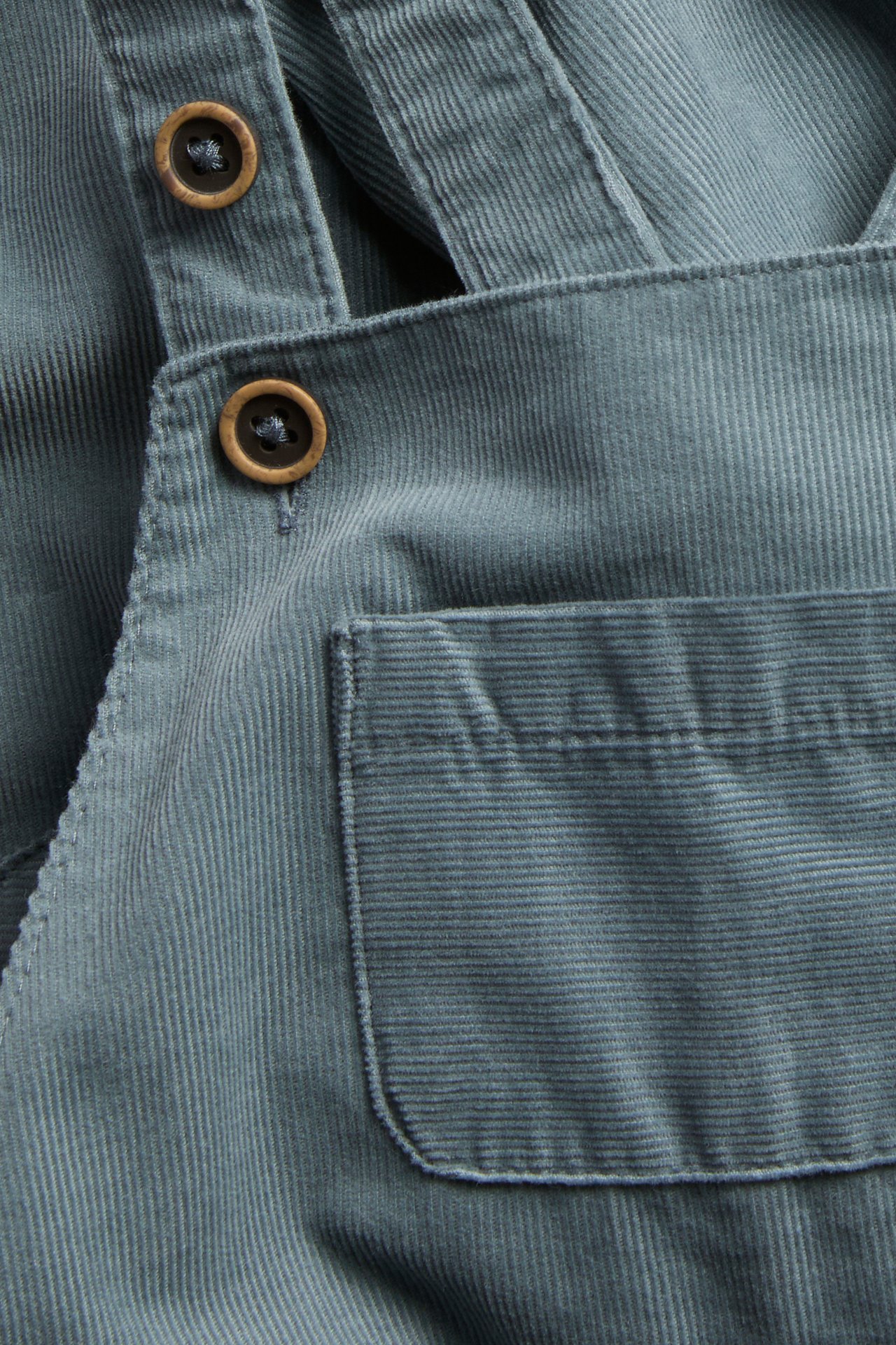 Nahaufnahme eines staubblauen Cord-Overalls mit zwei Holzknöpfen und einer Fronttasche.