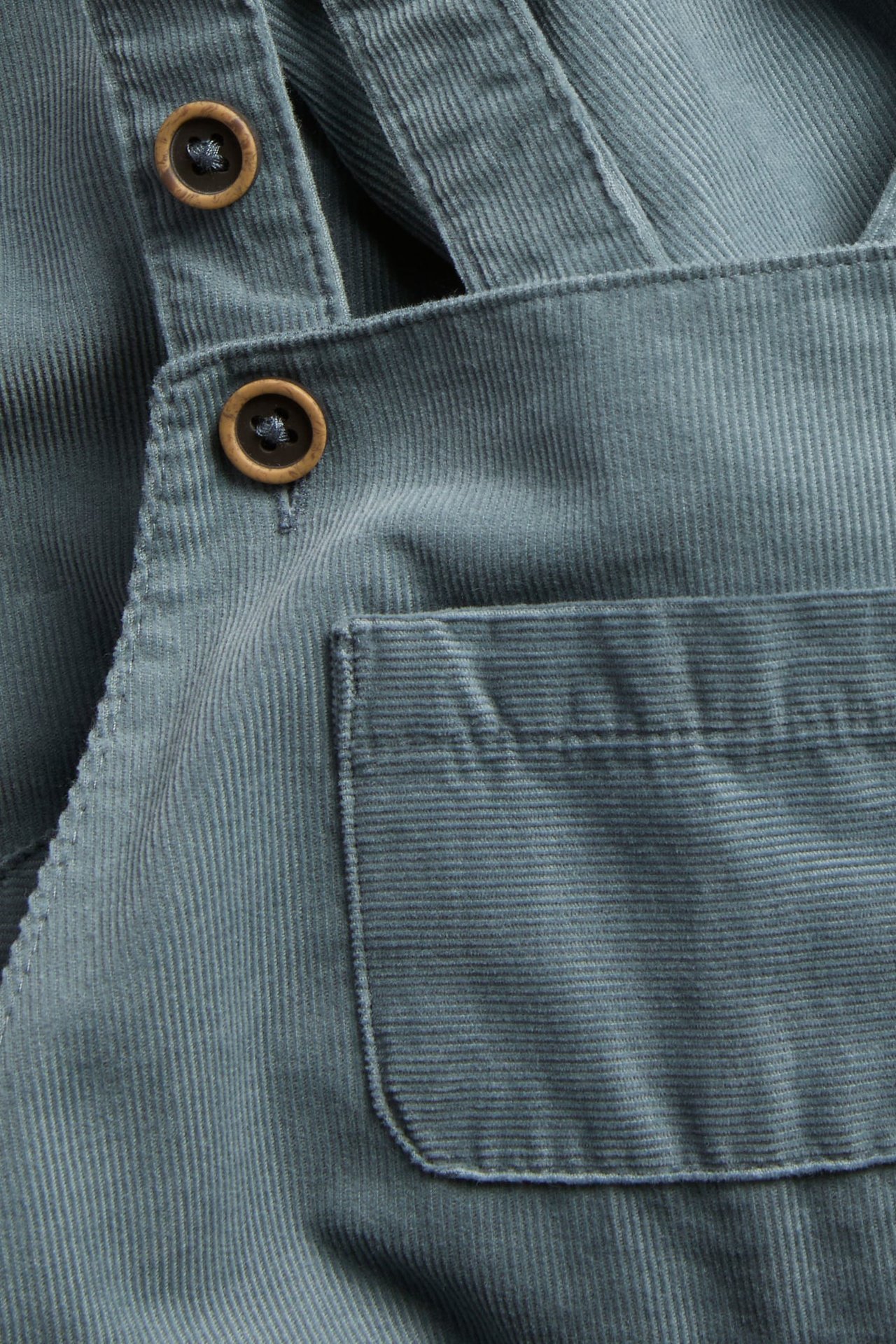 Nahaufnahme eines staubblauen Cord-Overalls mit zwei Holzknöpfen und einer Fronttasche.