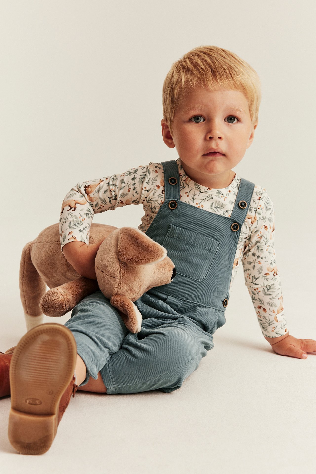 Kind in blauem Cord-Overall über weißem Langarmshirt mit Waldmuster, hält Plüschhund.