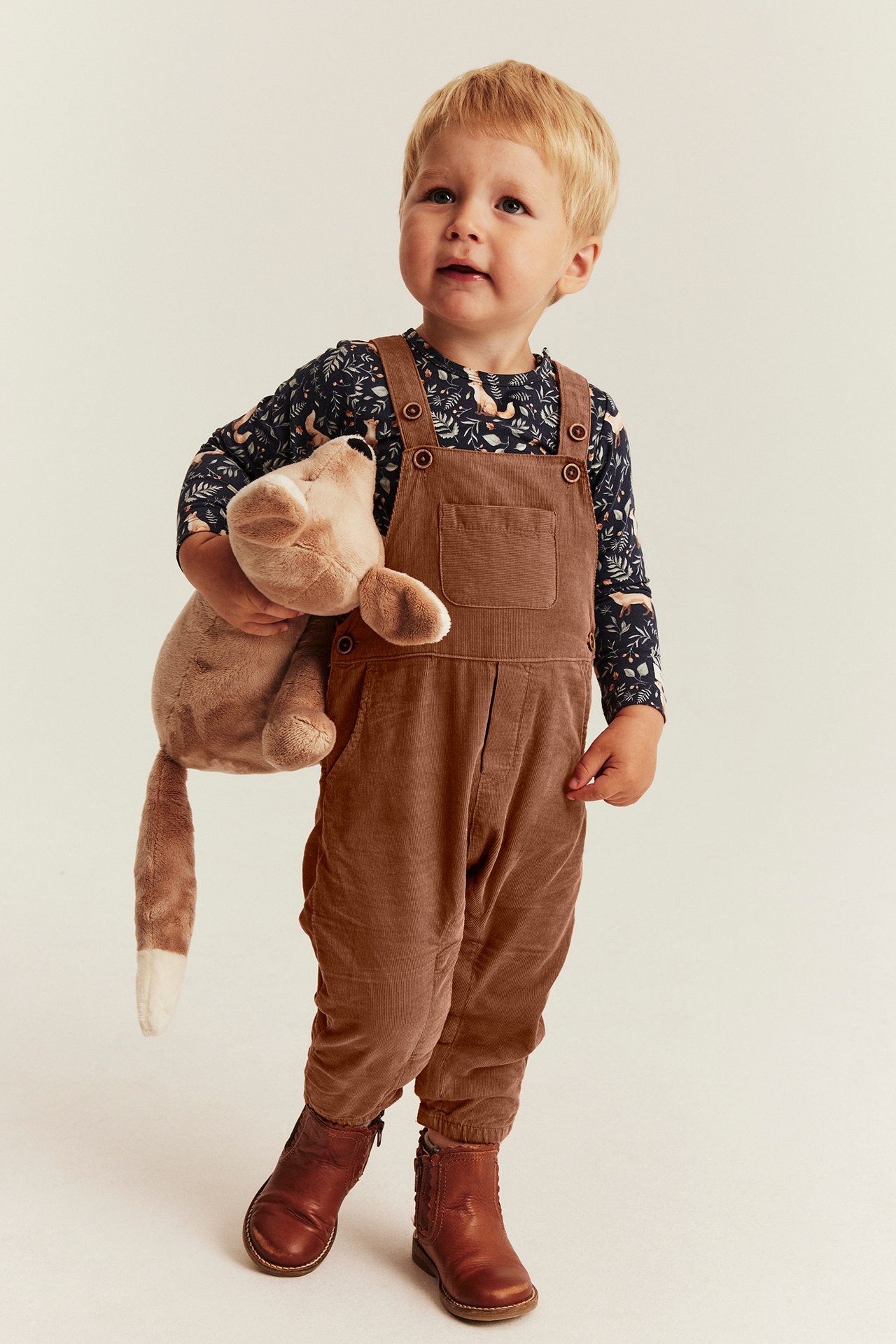 Front view of a kid in brown corduroy overalls, dark blue printed top, brown boots, holding a plush toy.