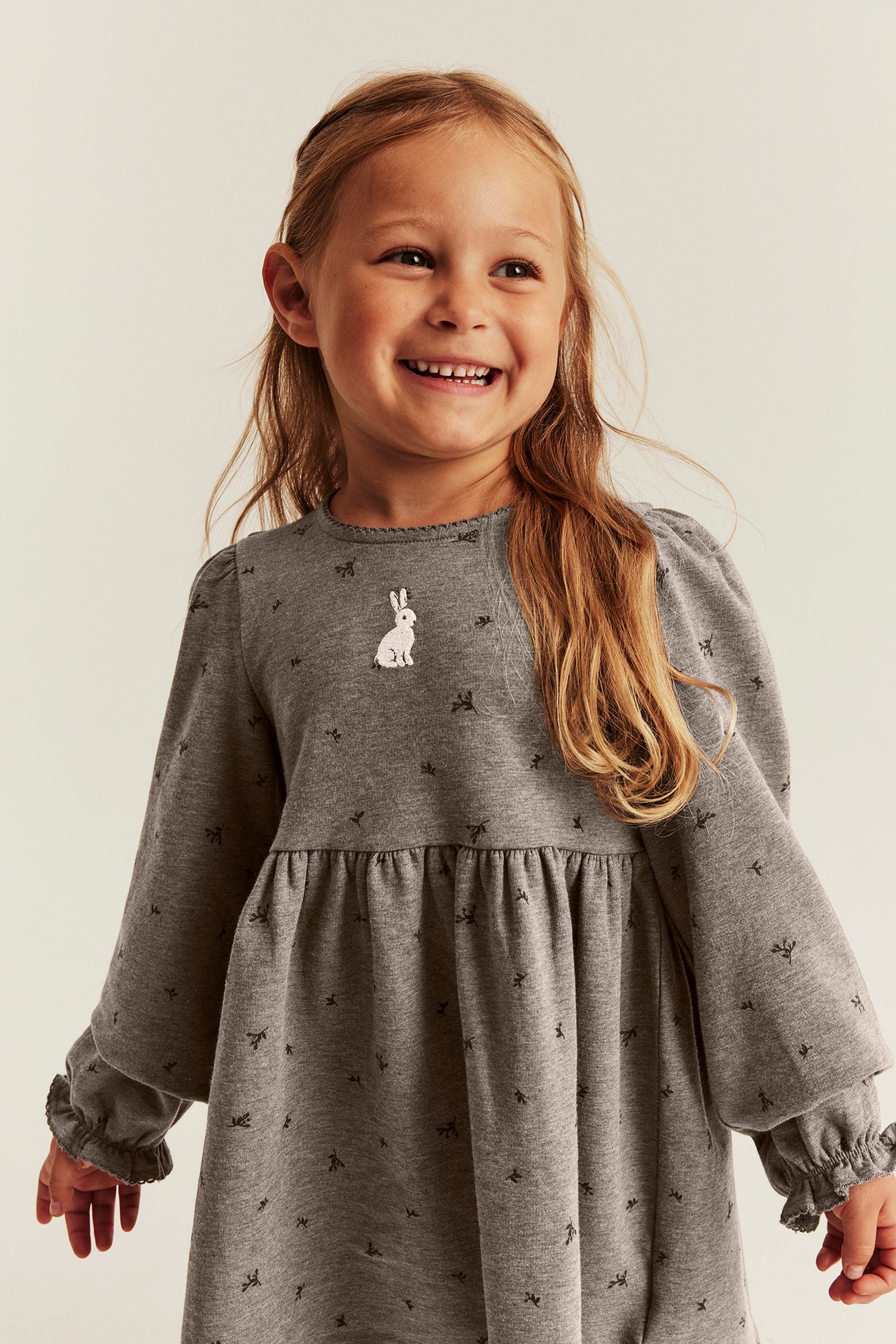 Lächelndes Kind in grauem Langarmkleid mit Schleifenmuster und weißer Hasenstickerei.