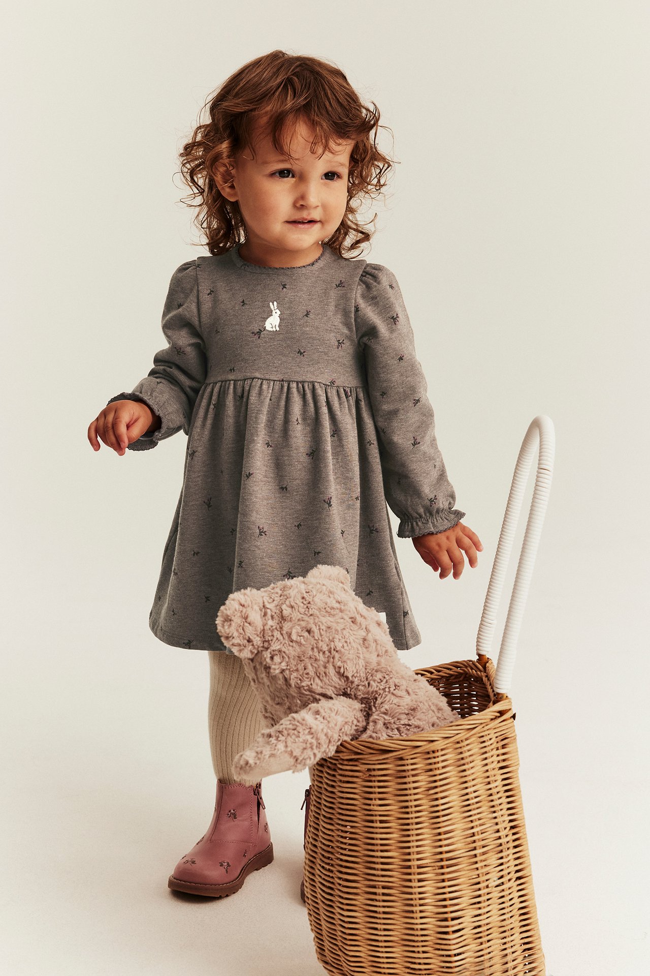 Vorderansicht: Kind trägt ein graues Langarmkleid mit weißem Hasen, rosa Stiefel und beigen Strumpfhosen.