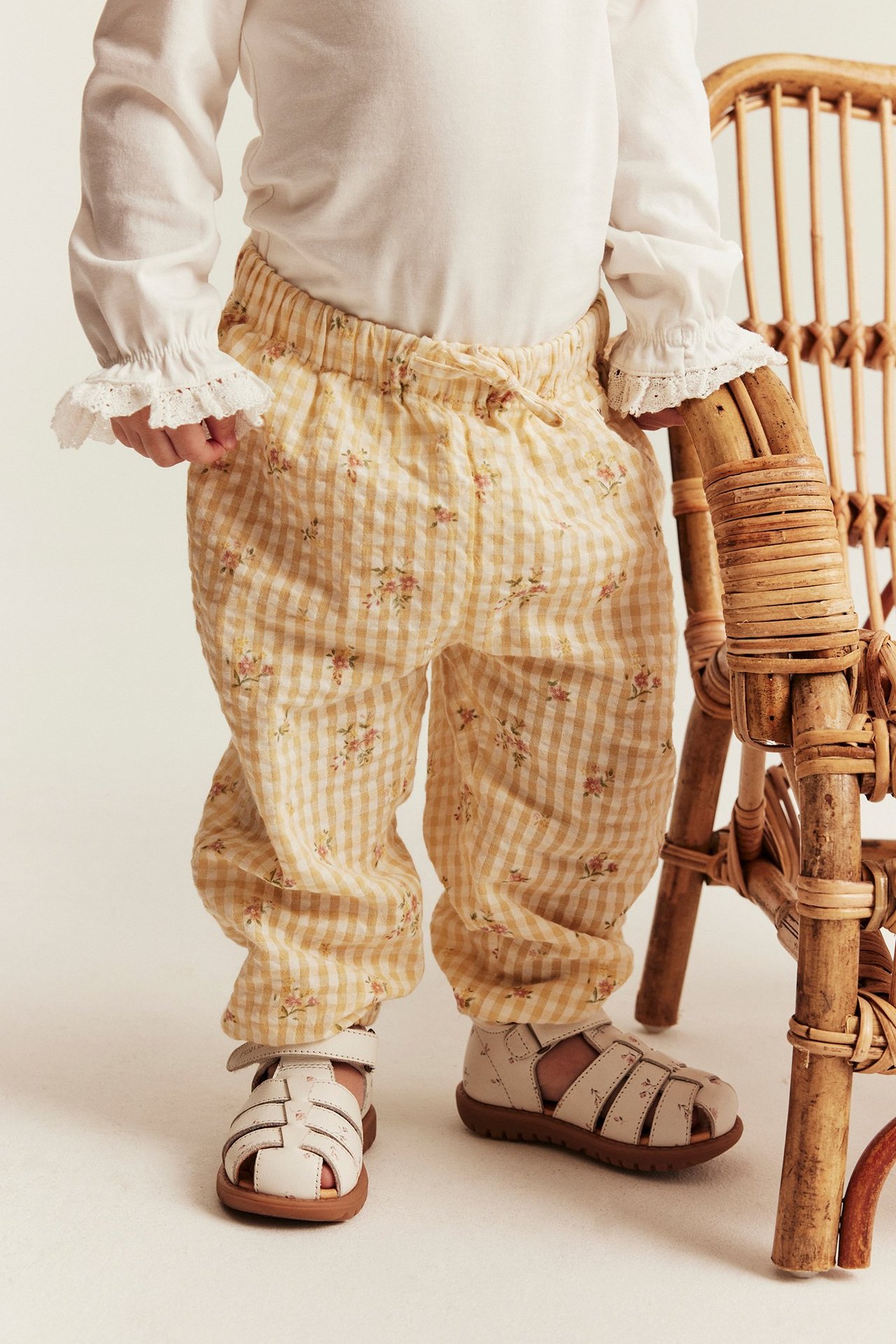 Vorderansicht: Ein Baby trägt gelbe karierte geblümte gewebte Baumwollhose, ein weißes Rüschenoberteil und beige Sandalen.