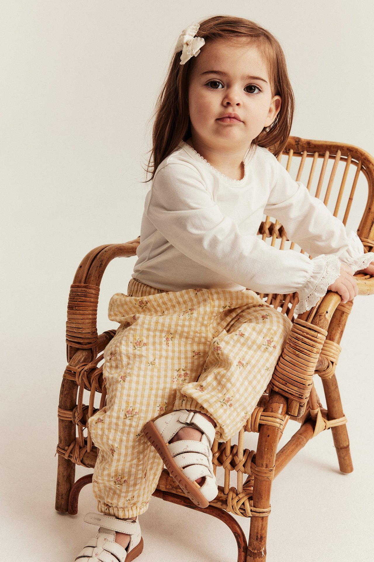 Kind trägt gelbe geblümte karierte Webhose, weißes Oberteil, weiße Sandalen und eine weiße Haarspange.