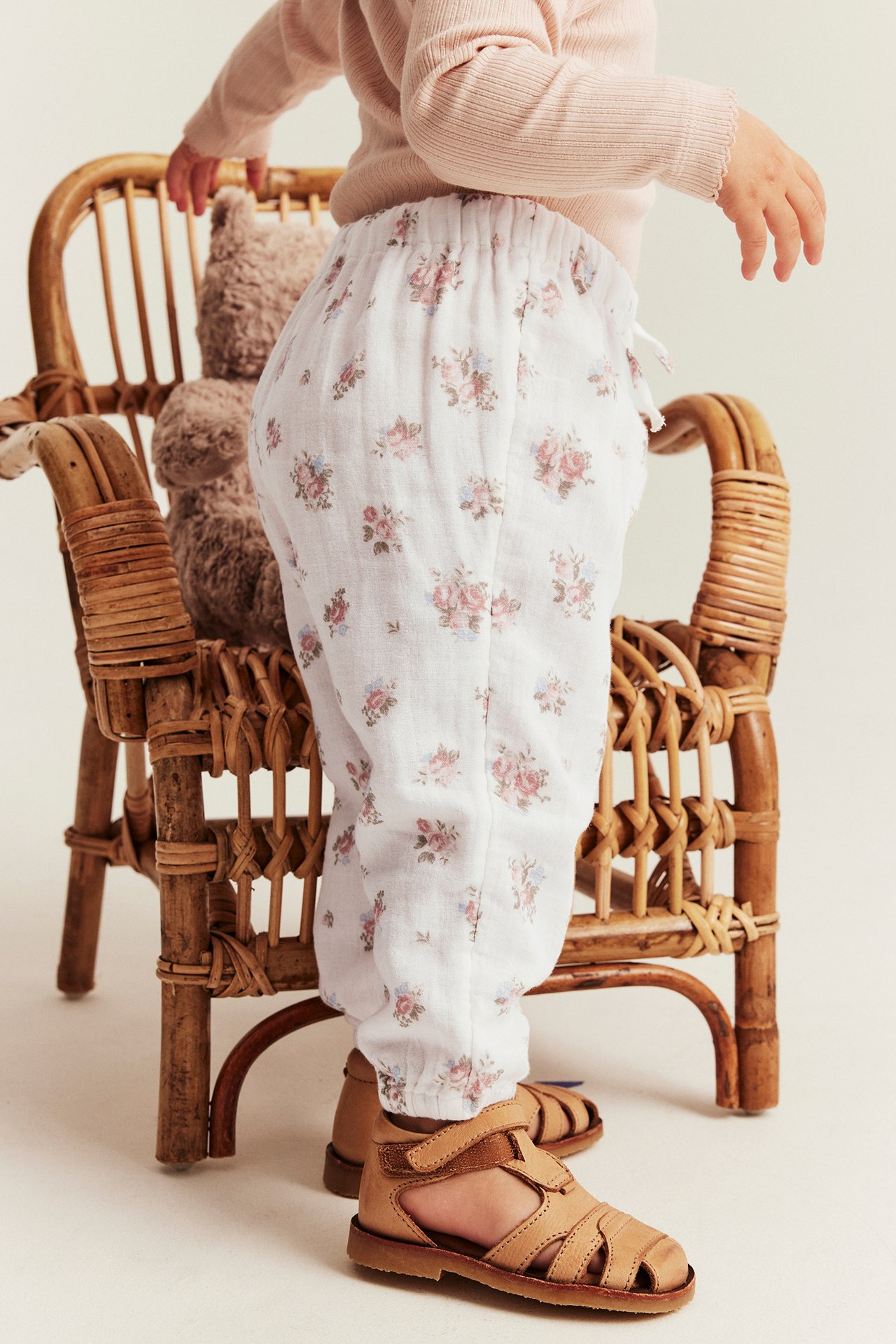 Side view: kid wears off-white floral cotton trousers, a pink long-sleeve top, and tan sandals.