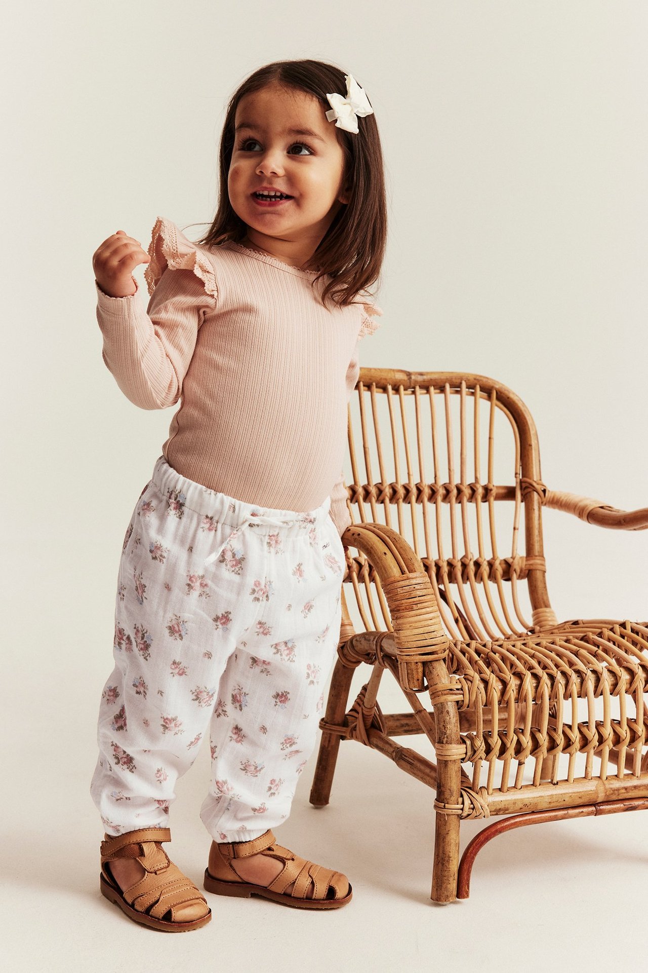 Front view: Kid wears offwhite floral woven cotton trousers, pink ribbed bodysuit, brown sandals, and an offwhite hair bow.