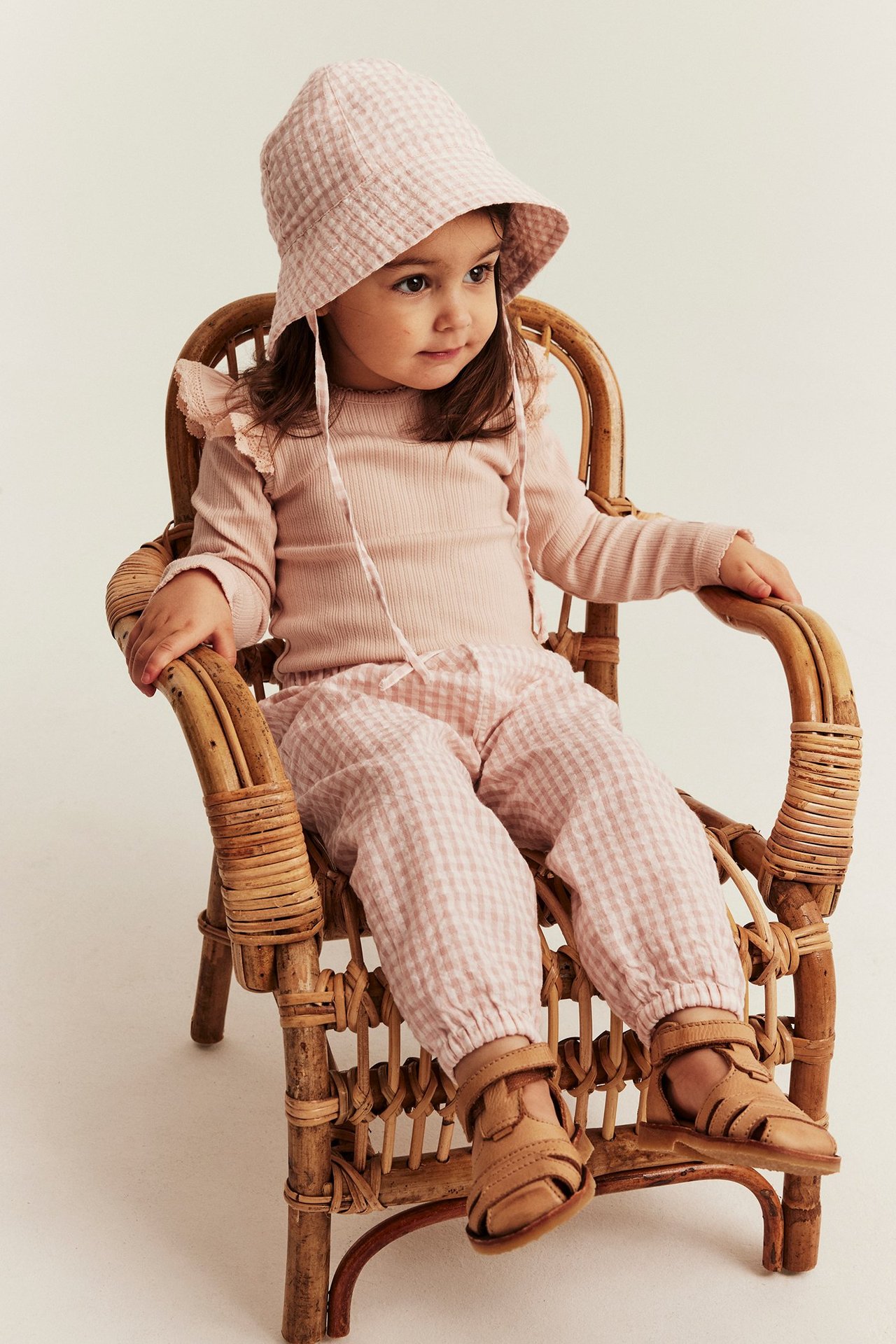 Vorderansicht: Kind trägt hellrosafarbene karierte gewebte Hose, ein Rippstricktop, passenden Sonnenhut und braune Sandalen.