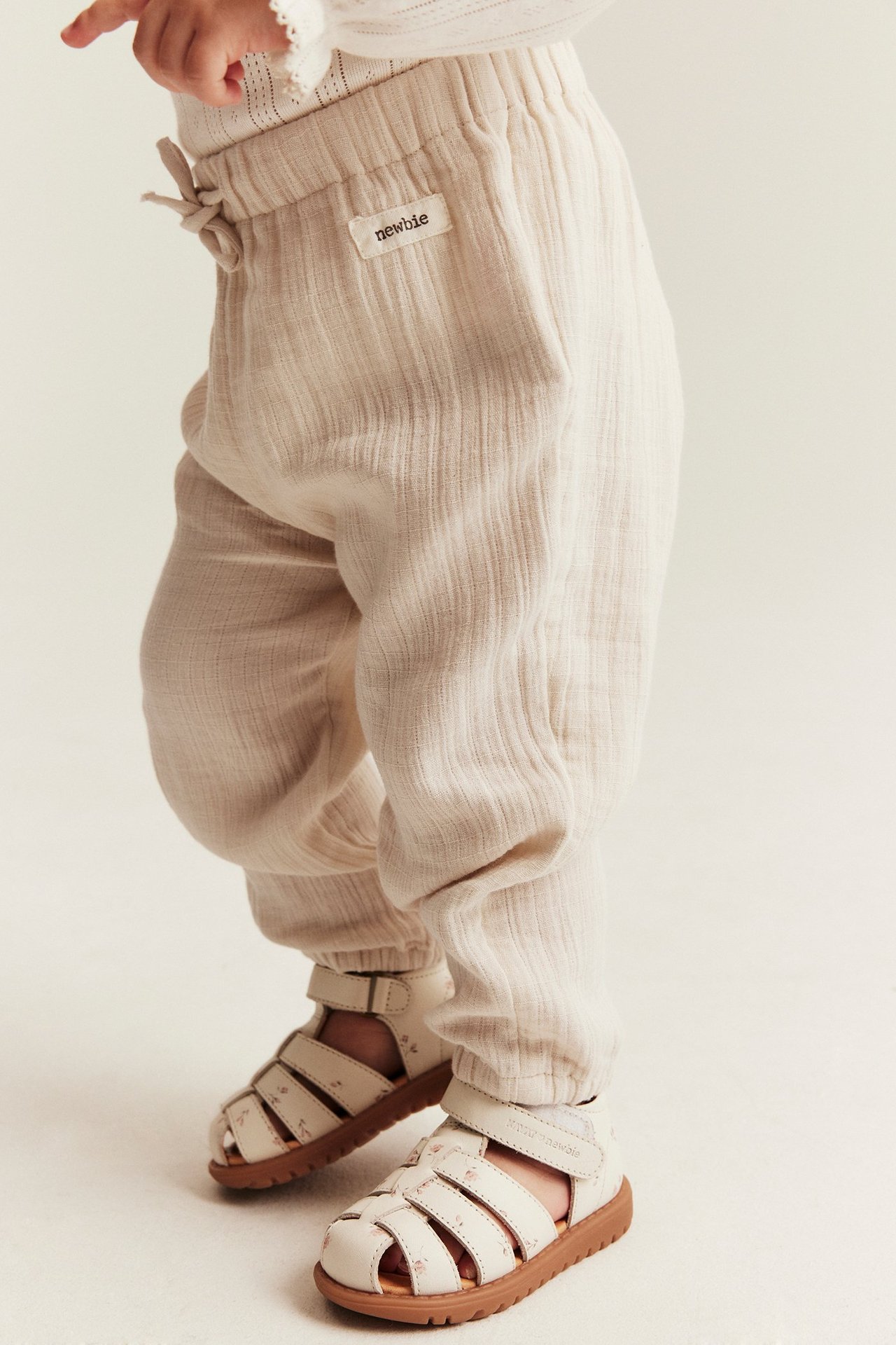 Seitenansicht eines Babys in hellbeiger gewebter Baumwollhose mit elastischem Bund und weißen gemusterten Sandalen.