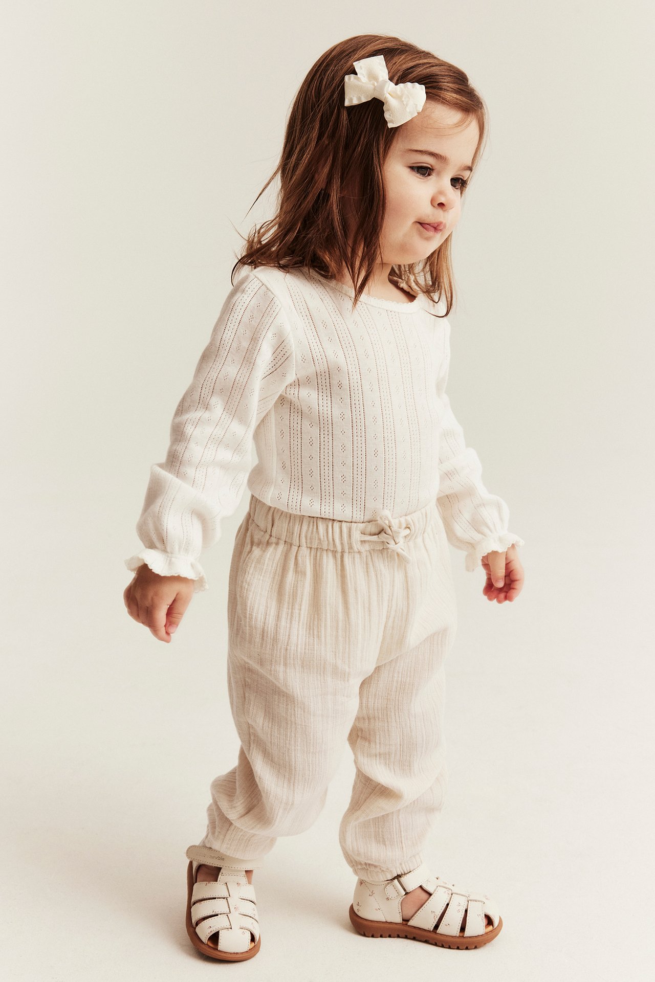 Front-side view: Kid wears light beige woven trousers with drawstring waist, white top, cream sandals, and a bow.