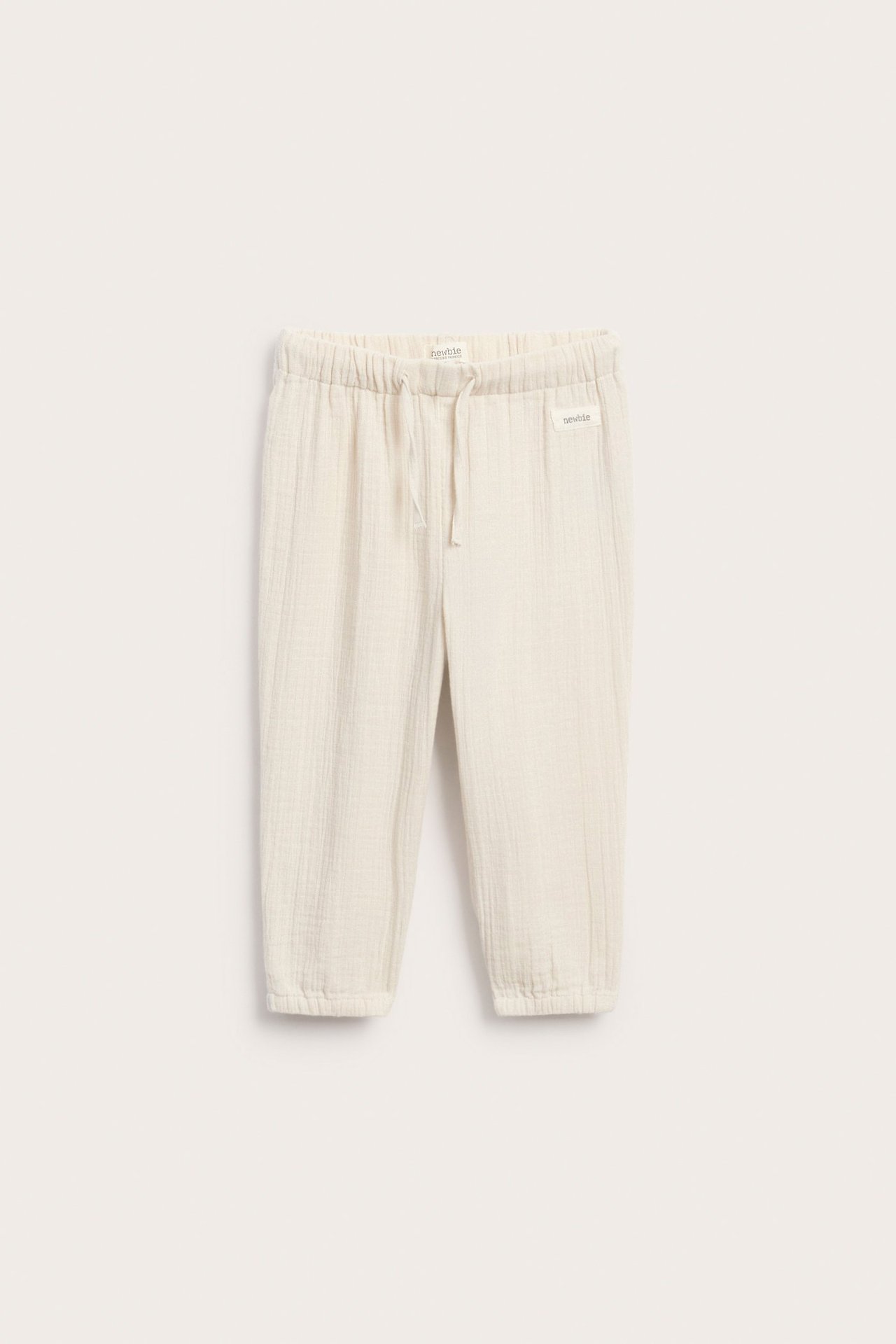 Front view of light beige woven cotton baby trousers with elastic waist, drawstring, and elasticated cuffs.