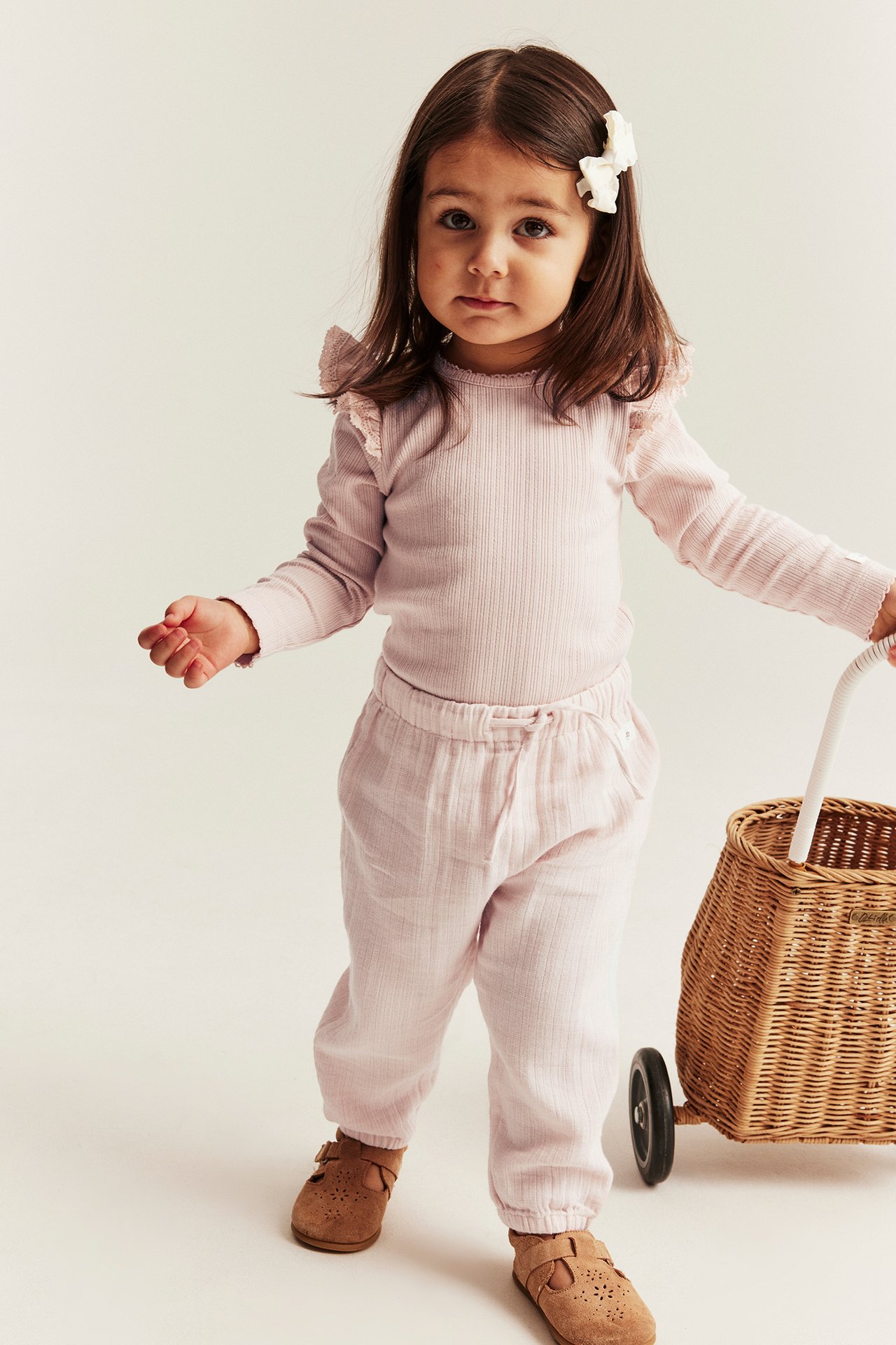 Front view: Kid in light pink woven cotton trousers, a light pink ribbed top, brown shoes, and a white bow.
