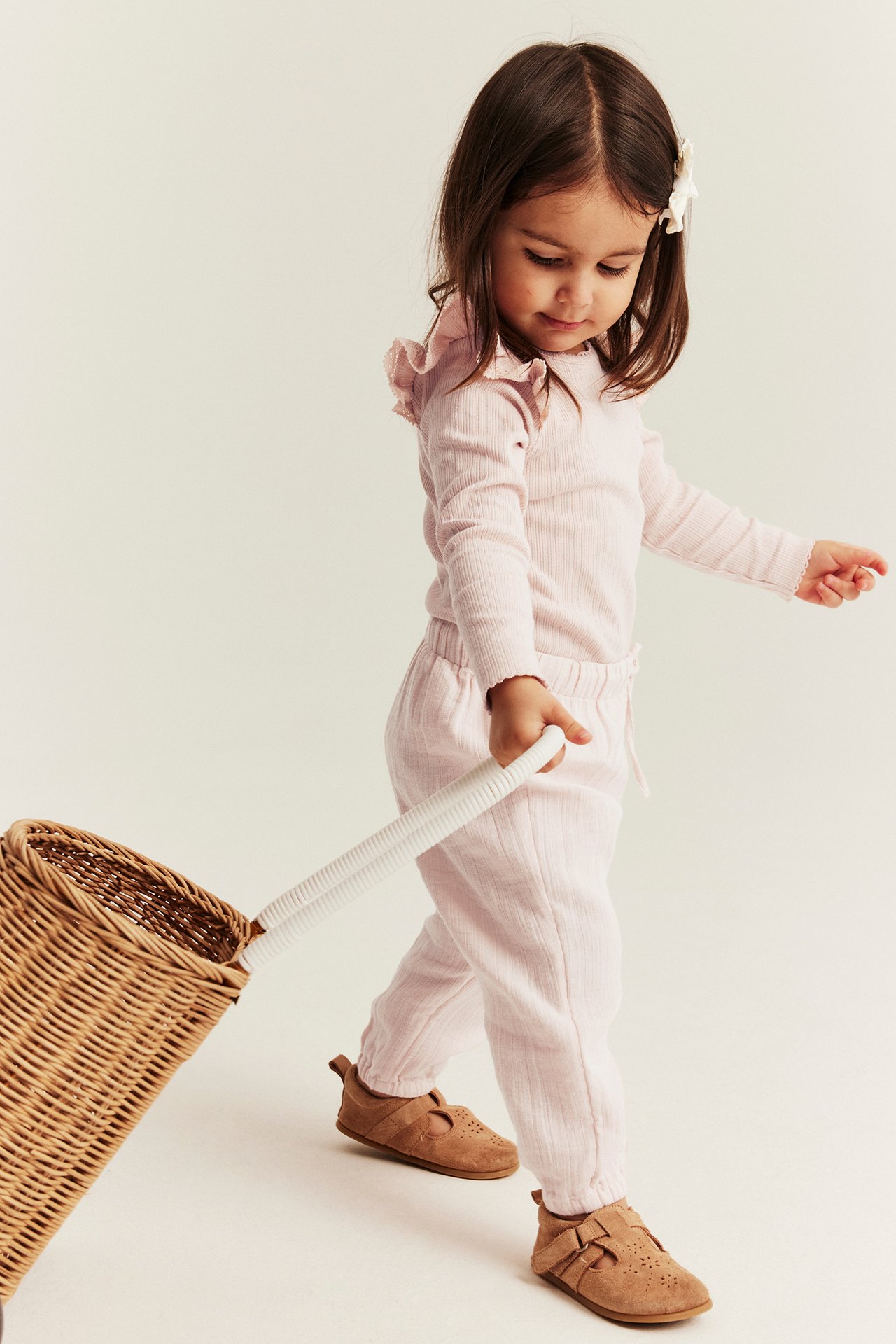 Side view of a kid in light pink woven trousers, a ruffled top, brown shoes, pulling a wicker basket.