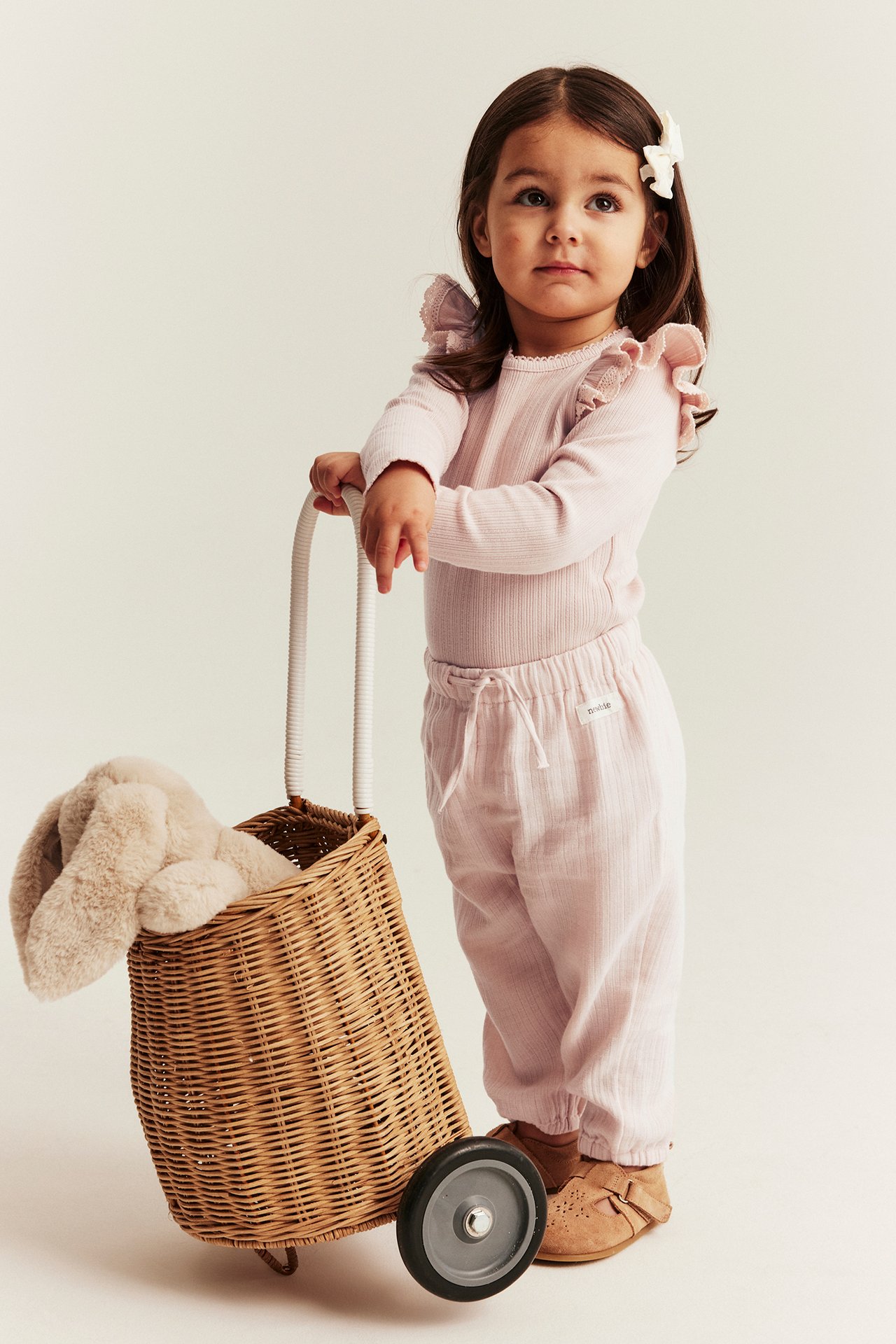 Kid in light pink woven pants with elastic waist, pink ribbed ruffle top, brown shoes, holding a toy basket.
