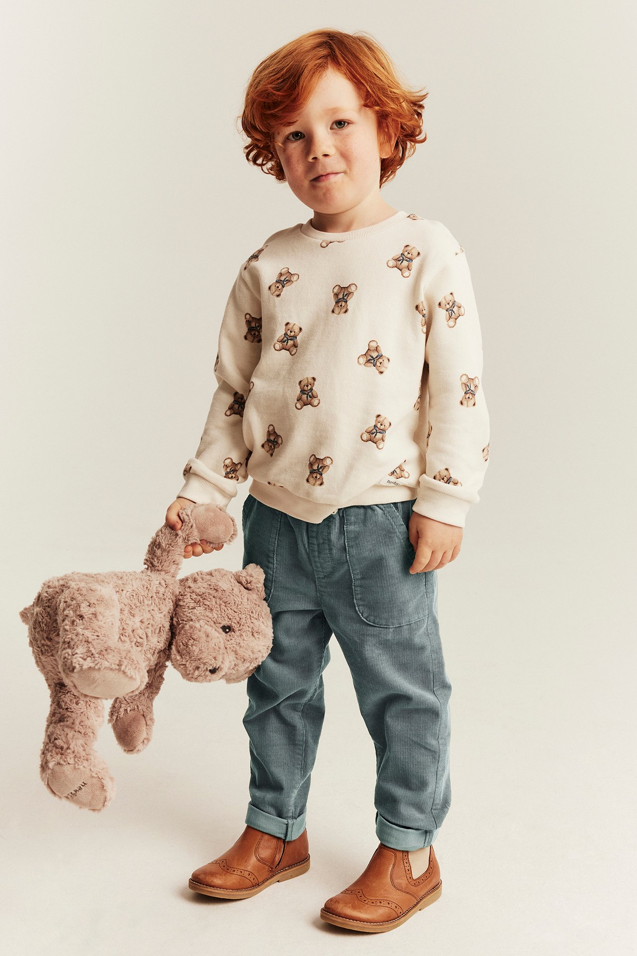 Vorderansicht: Kind trägt cremefarbenes Teddybär-Sweatshirt, blaue Cordhose, braune Schuhe, hält Plüschbär.
