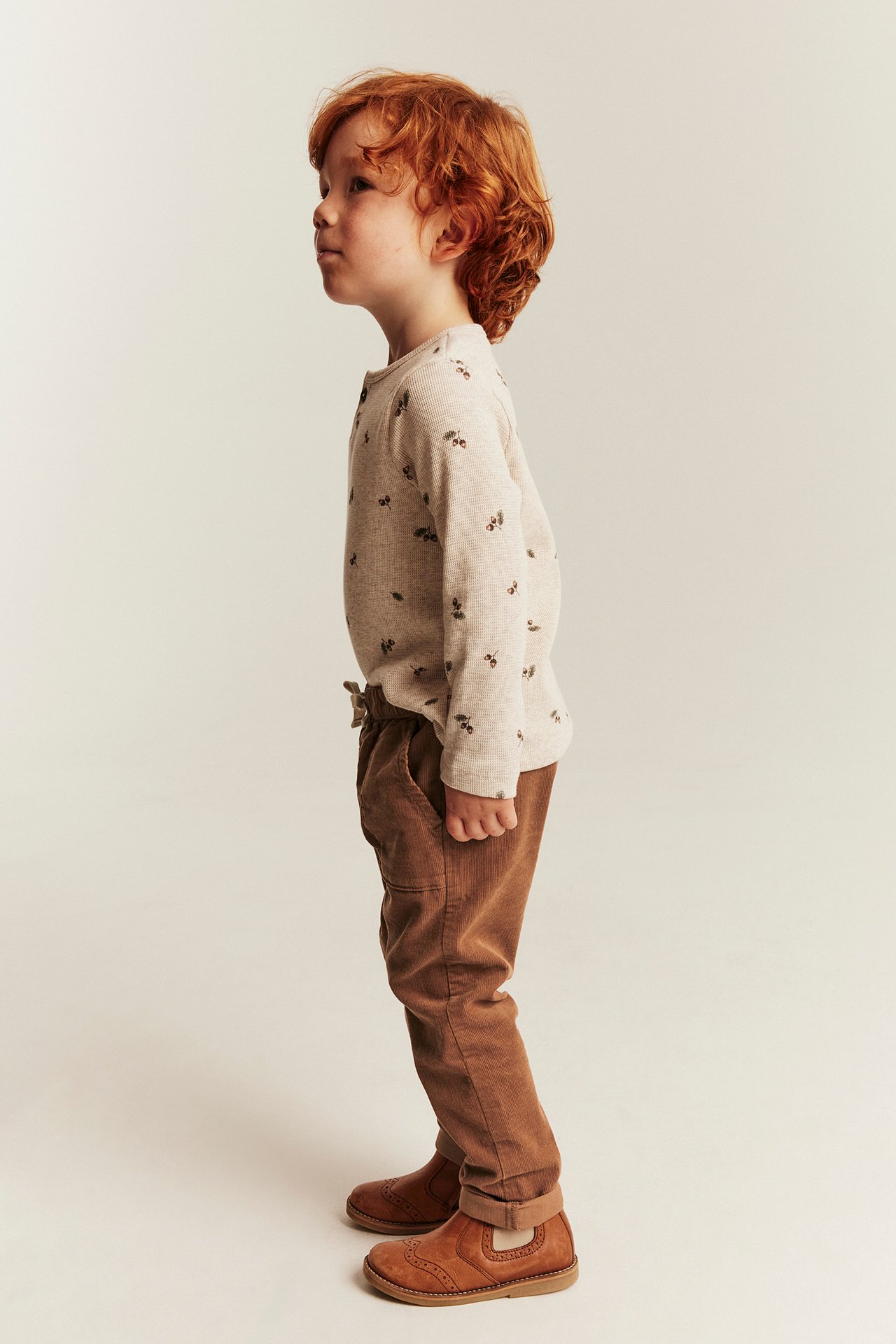 Kid in a beige ribbed long-sleeve top with dark print, brown corduroy pants, and brown boots.