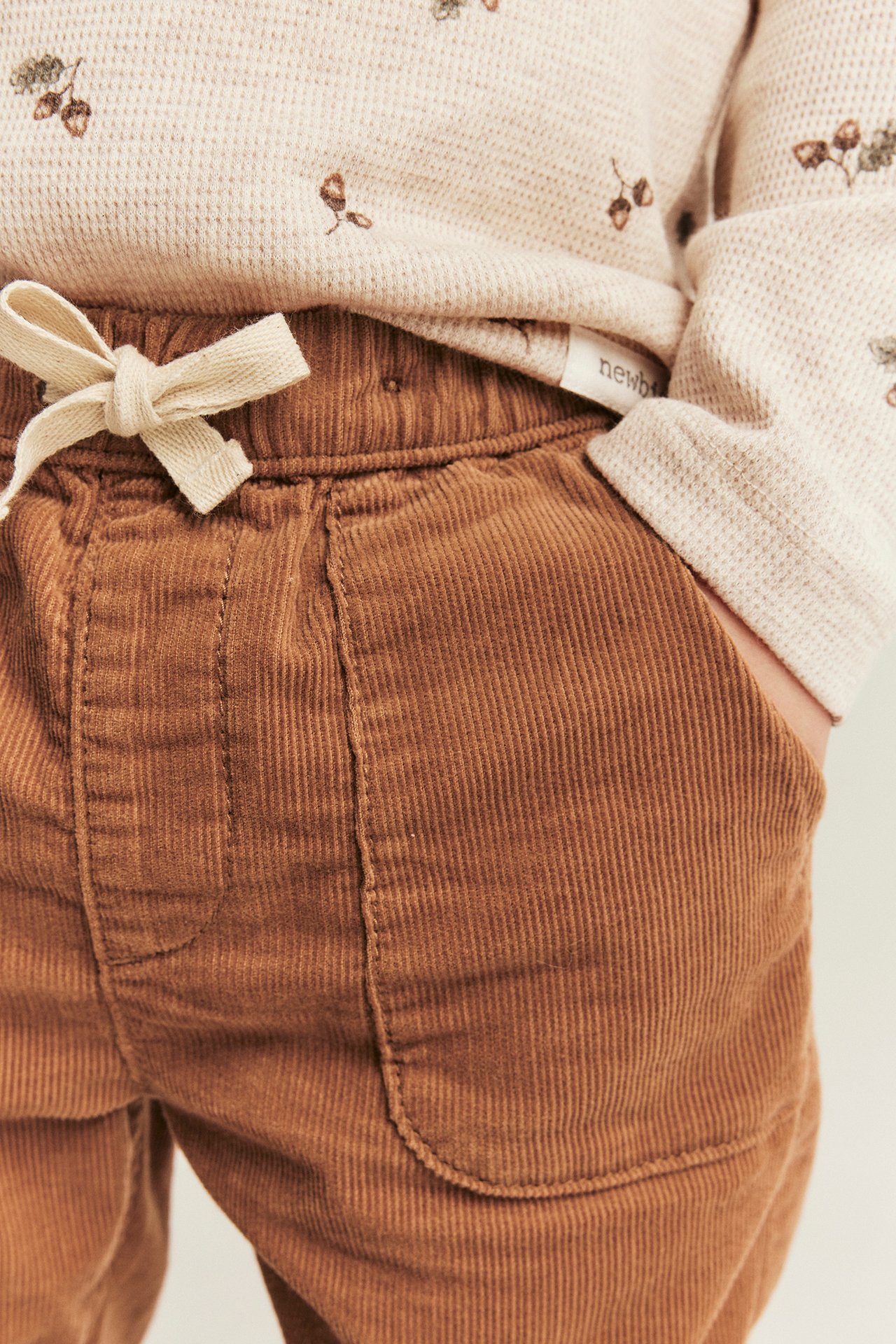 Nahaufnahme von Baby-Cordhose in Braun mit cremefarbenem Kordelzug und beigem gemustertem Waffelstrick-Oberteil.