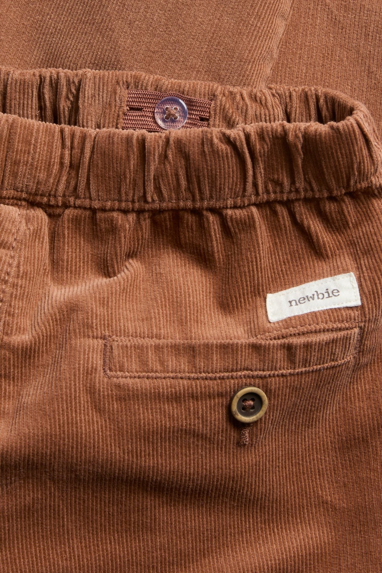 Nahaufnahme brauner Cordhose mit Gummizug, geknöpfter Gesäßtasche und weißem Etikett.