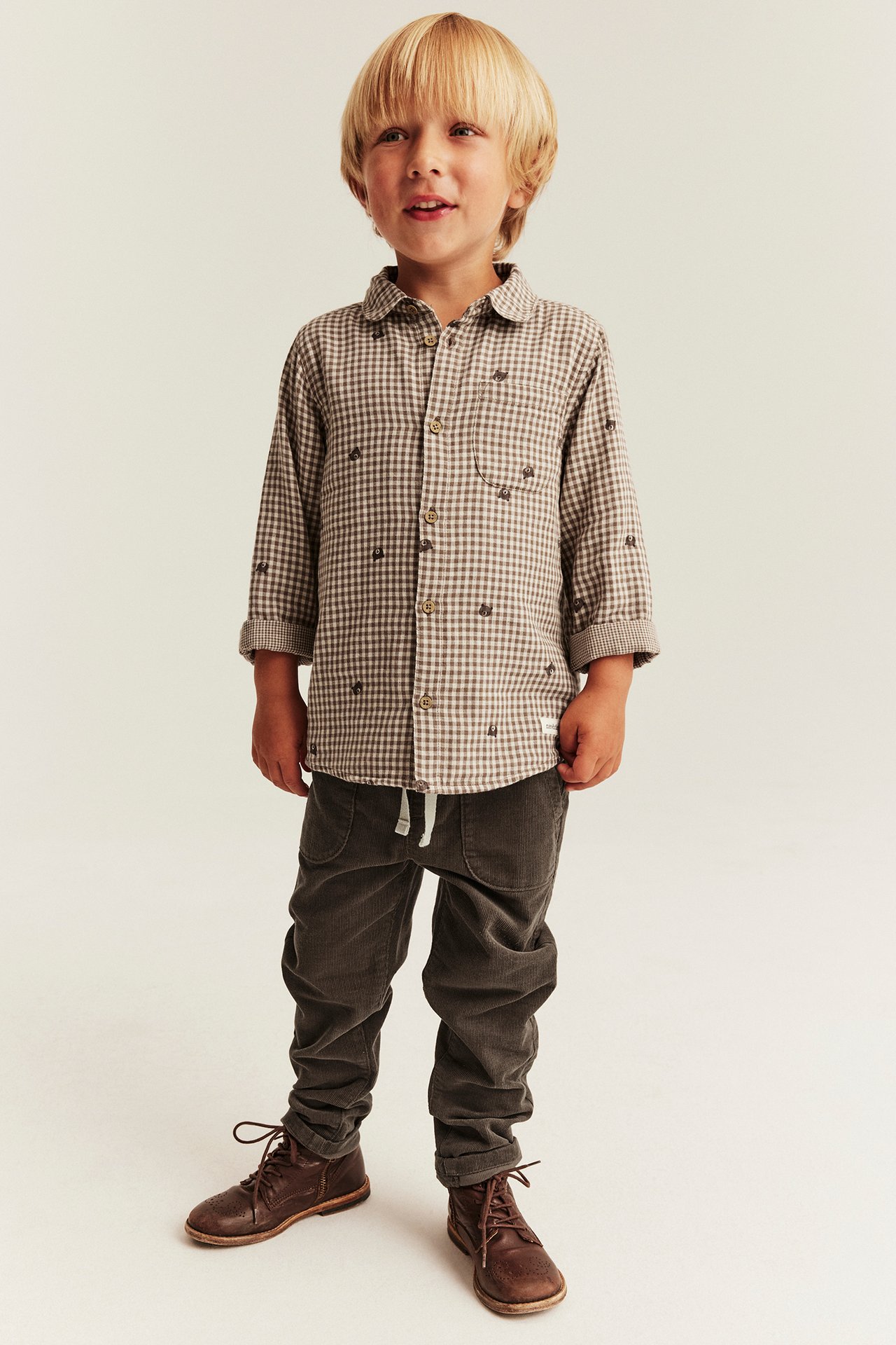 Kid in a brown checked shirt with embroideries, dark corduroy pants, and brown lace-up boots.