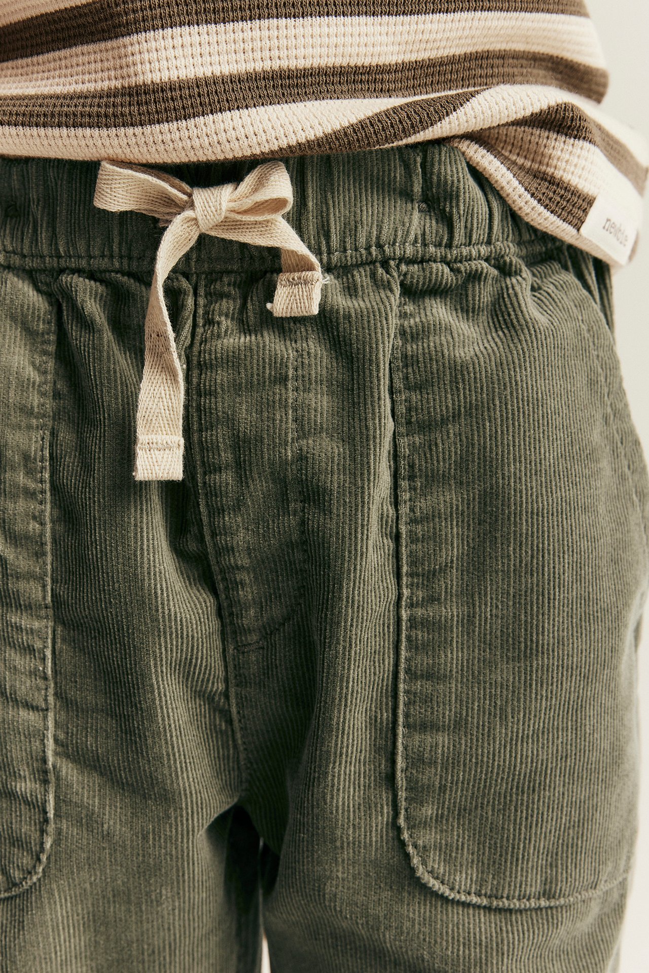 Nahaufnahme von Kinder-Cordhose in Grün mit Kordelzug und gestreiftem beigem Oberteil.
