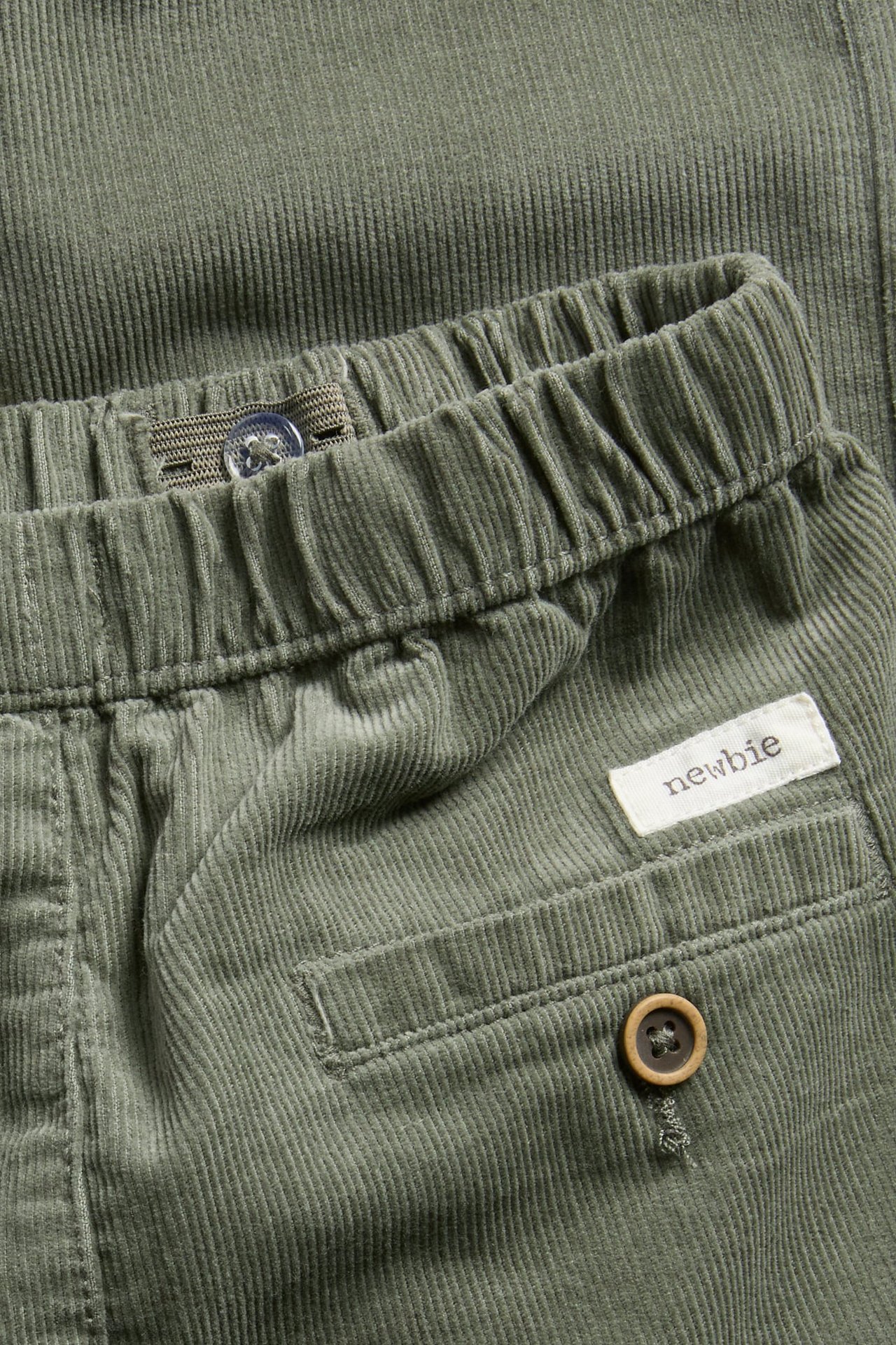 Nahaufnahme von grüner Cordhose mit elastischem Bund, innenliegender Knopfverstellung und geknöpfter Gesäßtasche.