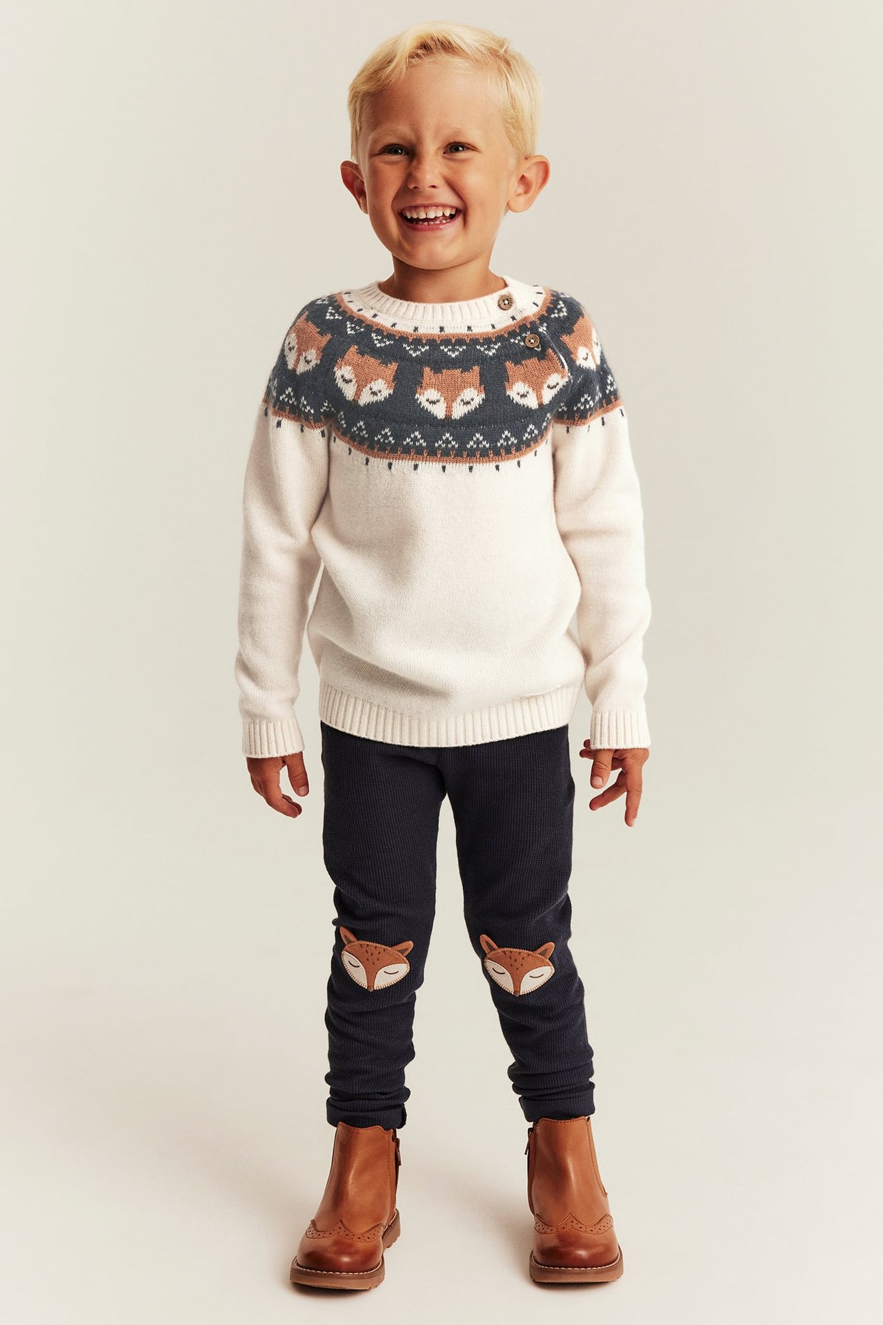 Vorderansicht eines Kindes in cremefarbenem Fuchs-Pullover, blauen Fuchs-Patch Leggings und braunen Stiefeln.