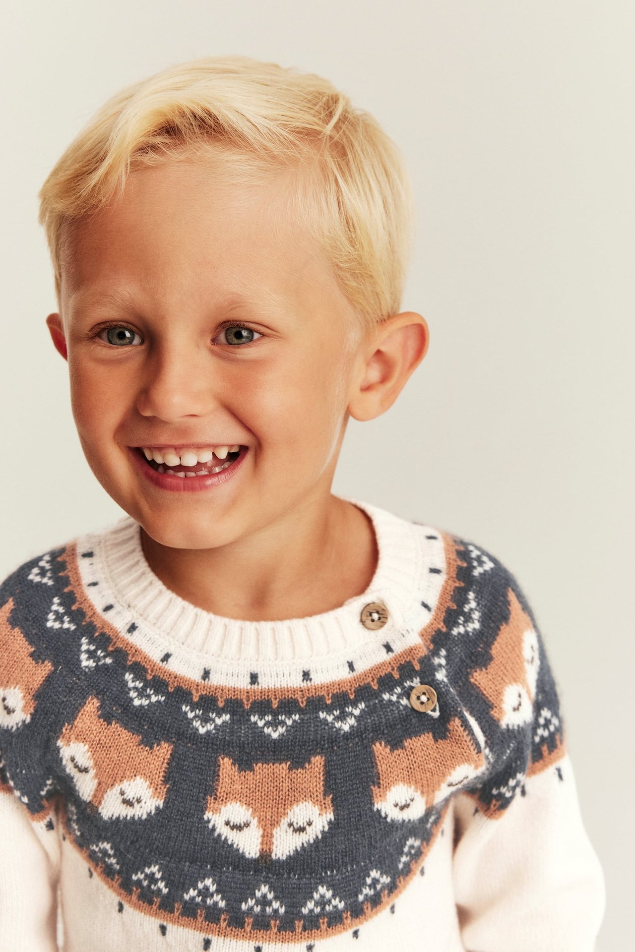 Nahaufnahme eines Kindes in einem cremefarbenen, grauen und orangefarbenen Pullover mit Fuchsmuster und Schulterknöpfen.