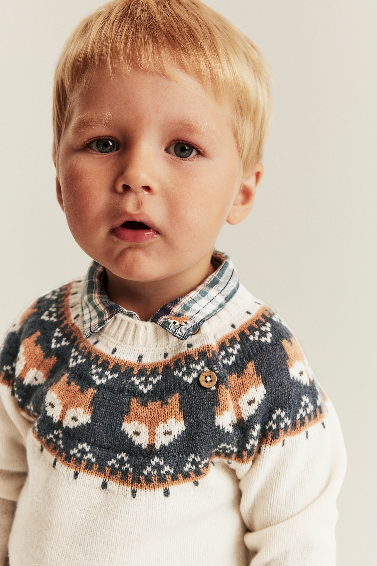 Vorderansicht: Babyjunge in cremefarbenem Strickpullover mit Fuchsmuster über kariertem Hemd.