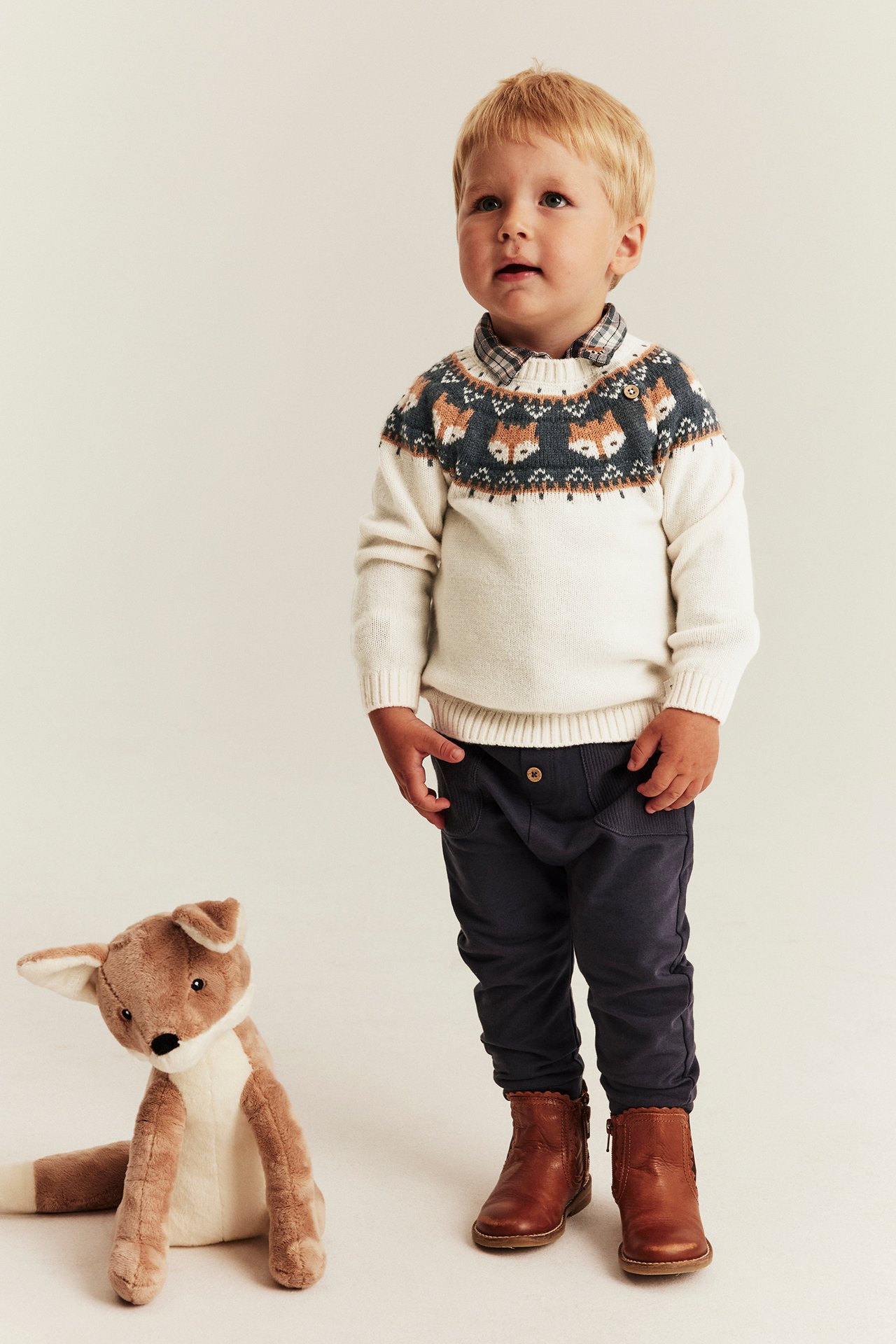 Babyjunge in cremefarbenem Fuchspullover, kariertem Hemd, dunkler Hose, braunen Stiefeln. Vorderansicht.