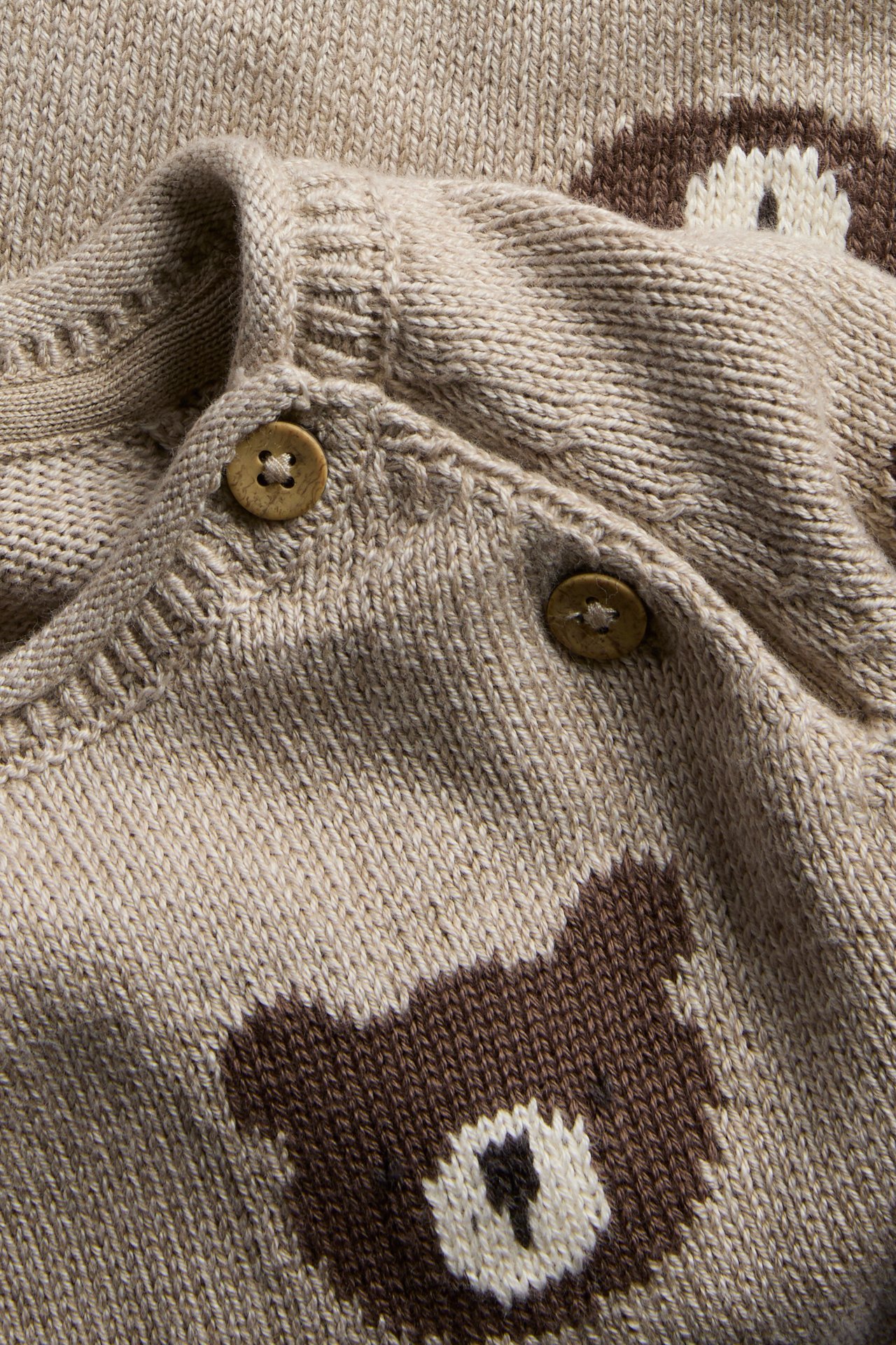 Nahaufnahme einer beigefarbenen gestrickten Baby-Strickjacke mit braunen Bärenmustern und Holzknöpfen.
