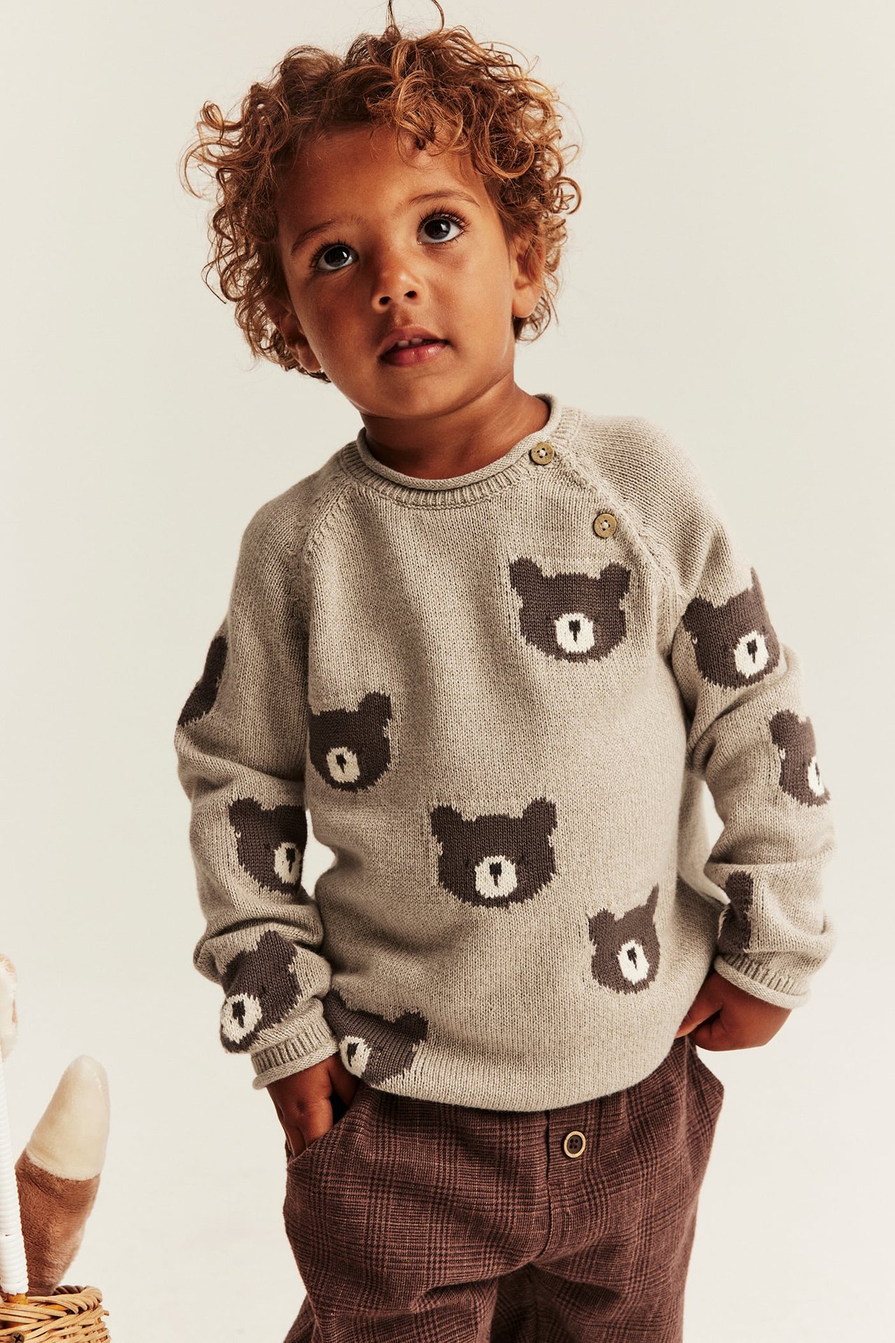 Frontansicht eines Kindes in beigefarbenem Strickpullover mit Bärenmuster und brauner Karohose.