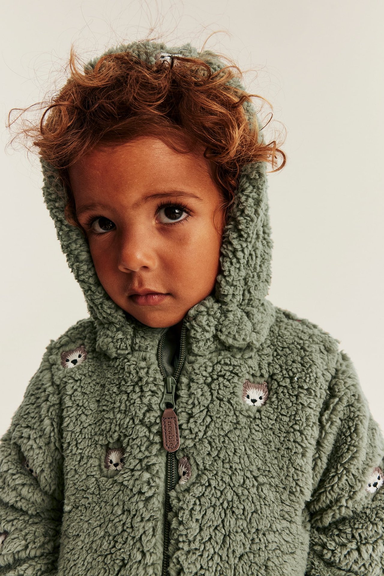 Close-up front view of a kid wearing a green hooded teddy jacket with hedgehog embroidery.