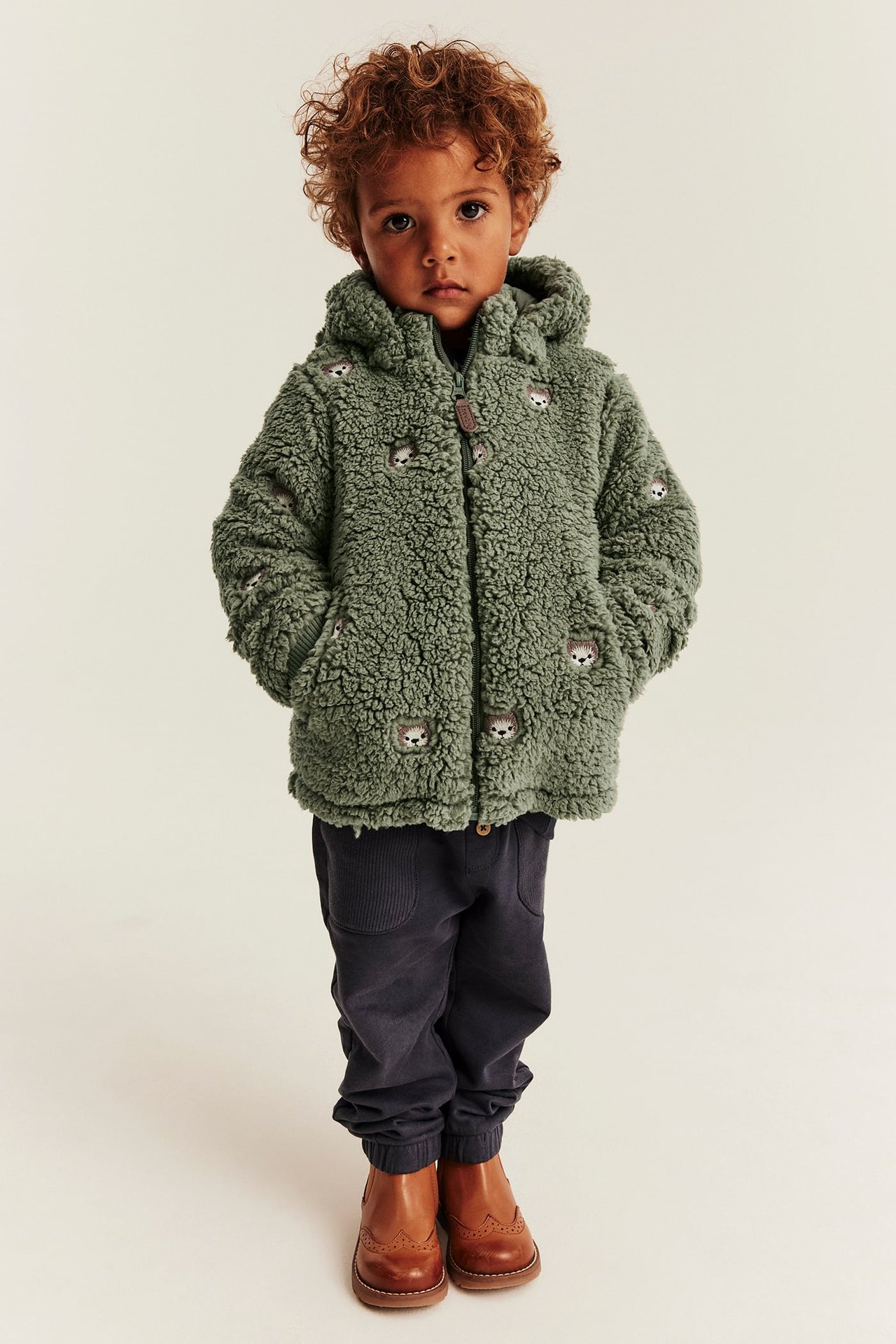 Front view: Kid wears green hedgehog embroidered hooded teddy jacket, dark blue corduroy pants, brown boots.
