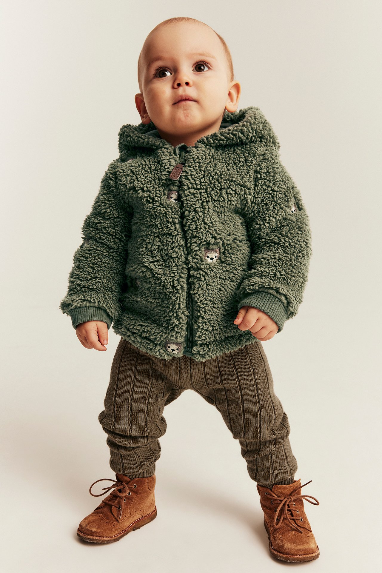 Baby in a green fluffy hooded jacket with hedgehog details, brown ribbed pants, and brown boots.