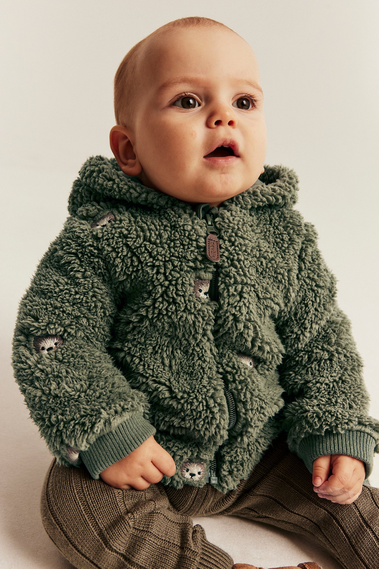 Baby in a green fluffy hooded jacket with hedgehog embroidery and brown ribbed pants.