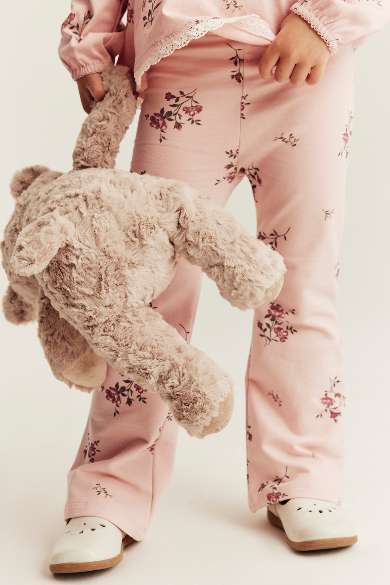 Kid wears light pink floral jersey flared pants, matching top, white shoes, and holds a brown teddy bear.