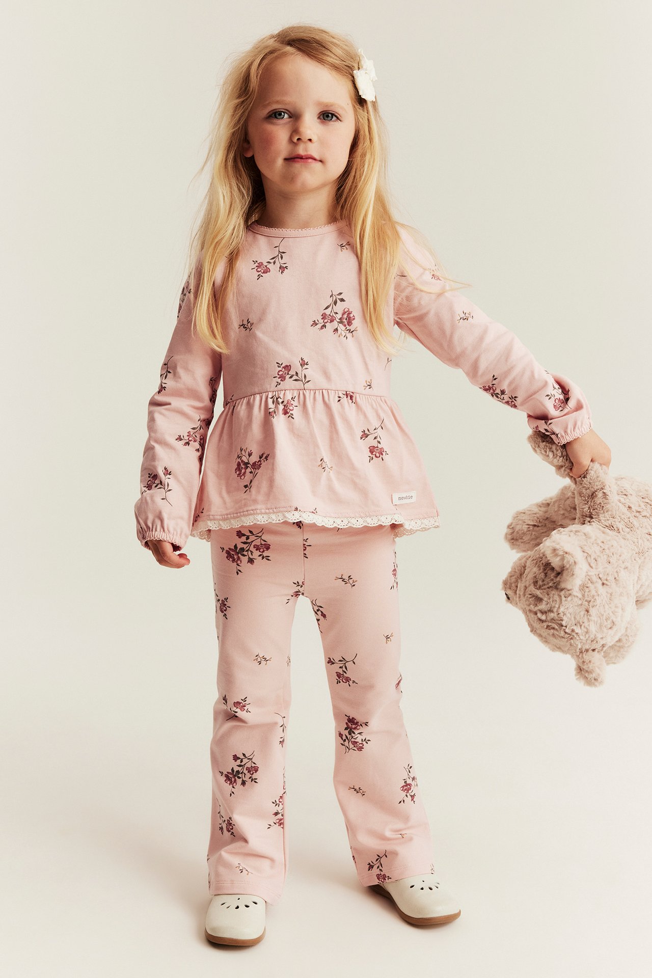 Front view of a kid wearing pink floral jersey flared pants and a matching peplum top. She holds a teddy bear.
