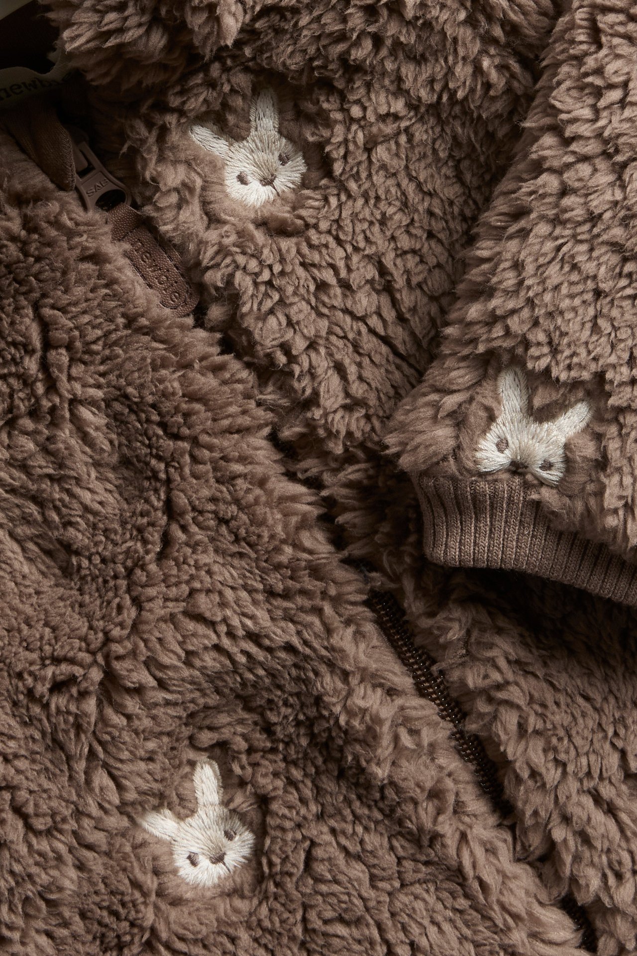 Close-up of a brown fluffy baby or kid's garment with white bunny embroidery and zipper.